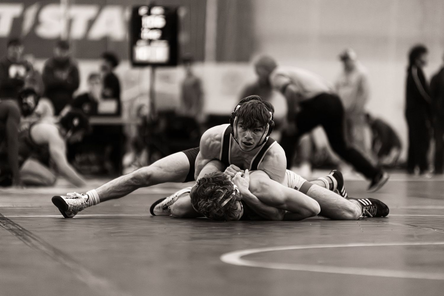 Two wrestlers compete on a mat during a match while others observe in the background.