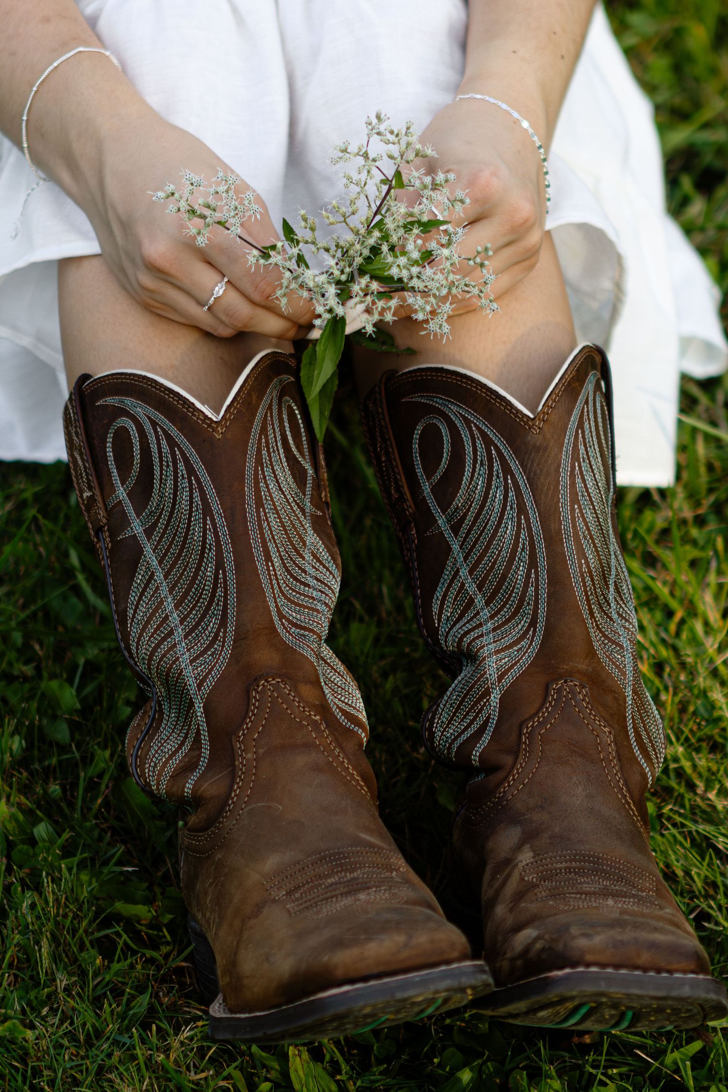 Brown leather cowboy boots with white floral details sit on green grass during an outdoor photoshoot.