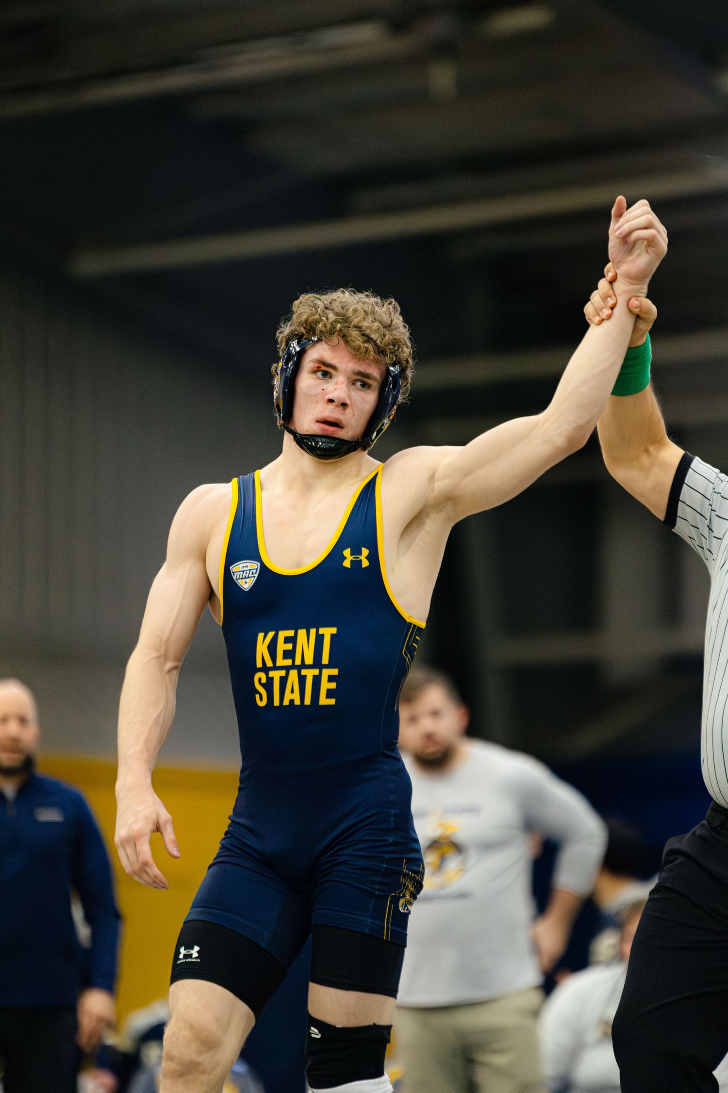 College wrestler in navy and gold uniform performs dynamic move during competitive match on wrestling mat.