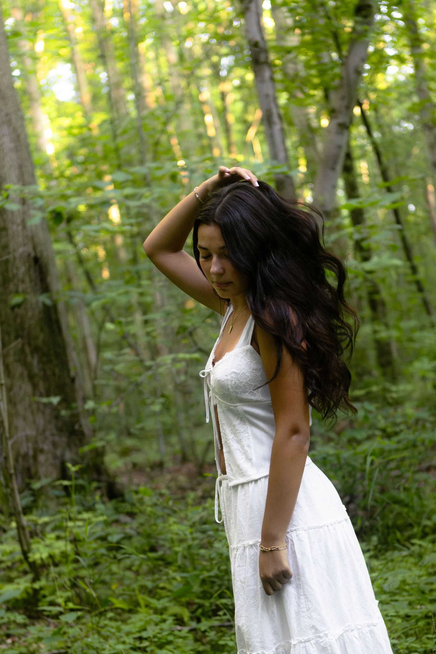 A figure in a flowing white summer dress poses expressively among lush green forest trees in natural light.