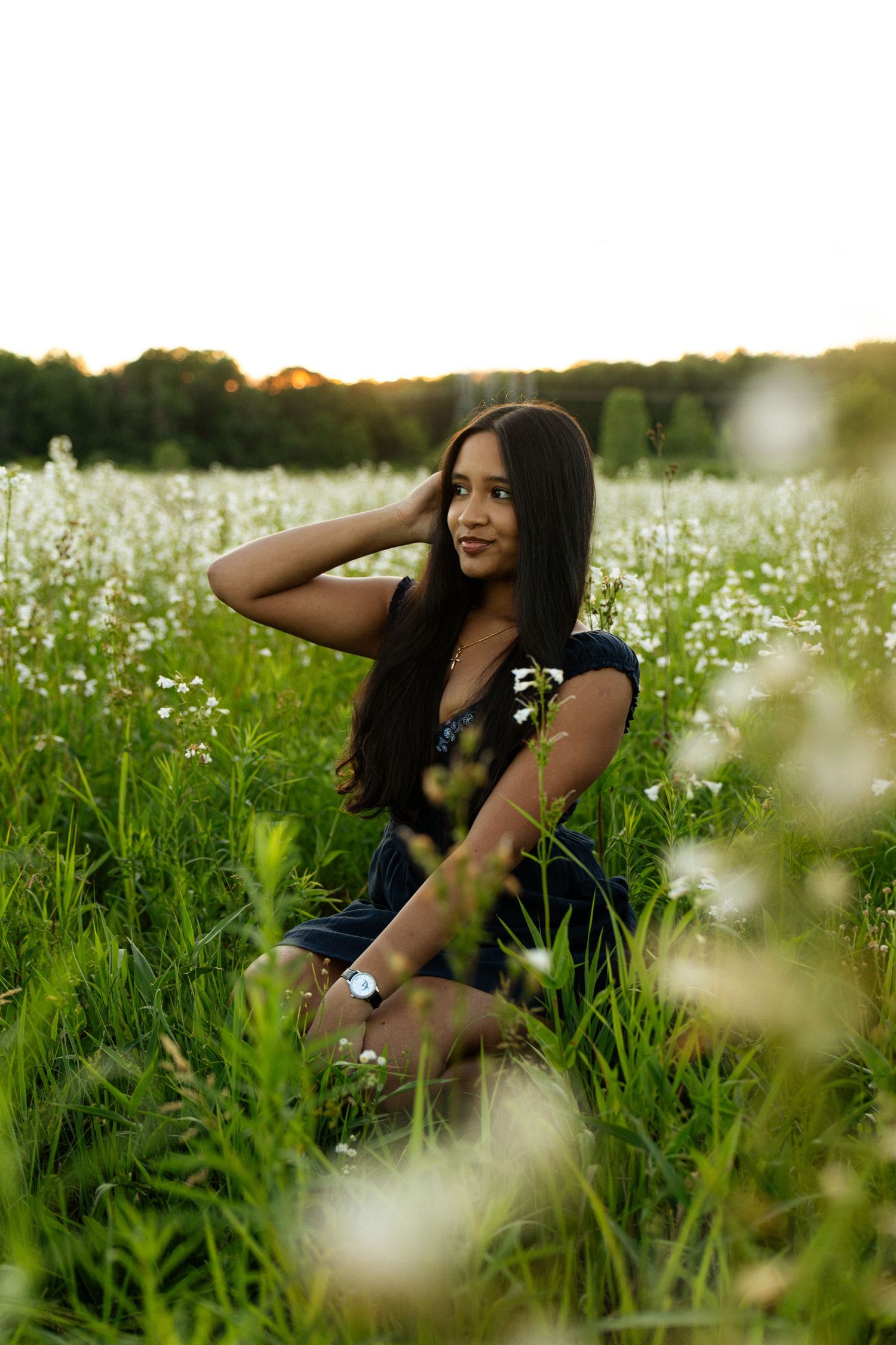 Young woman with long dark hair sits gracefully in a lush green field of white wildflowers at golden sunset.