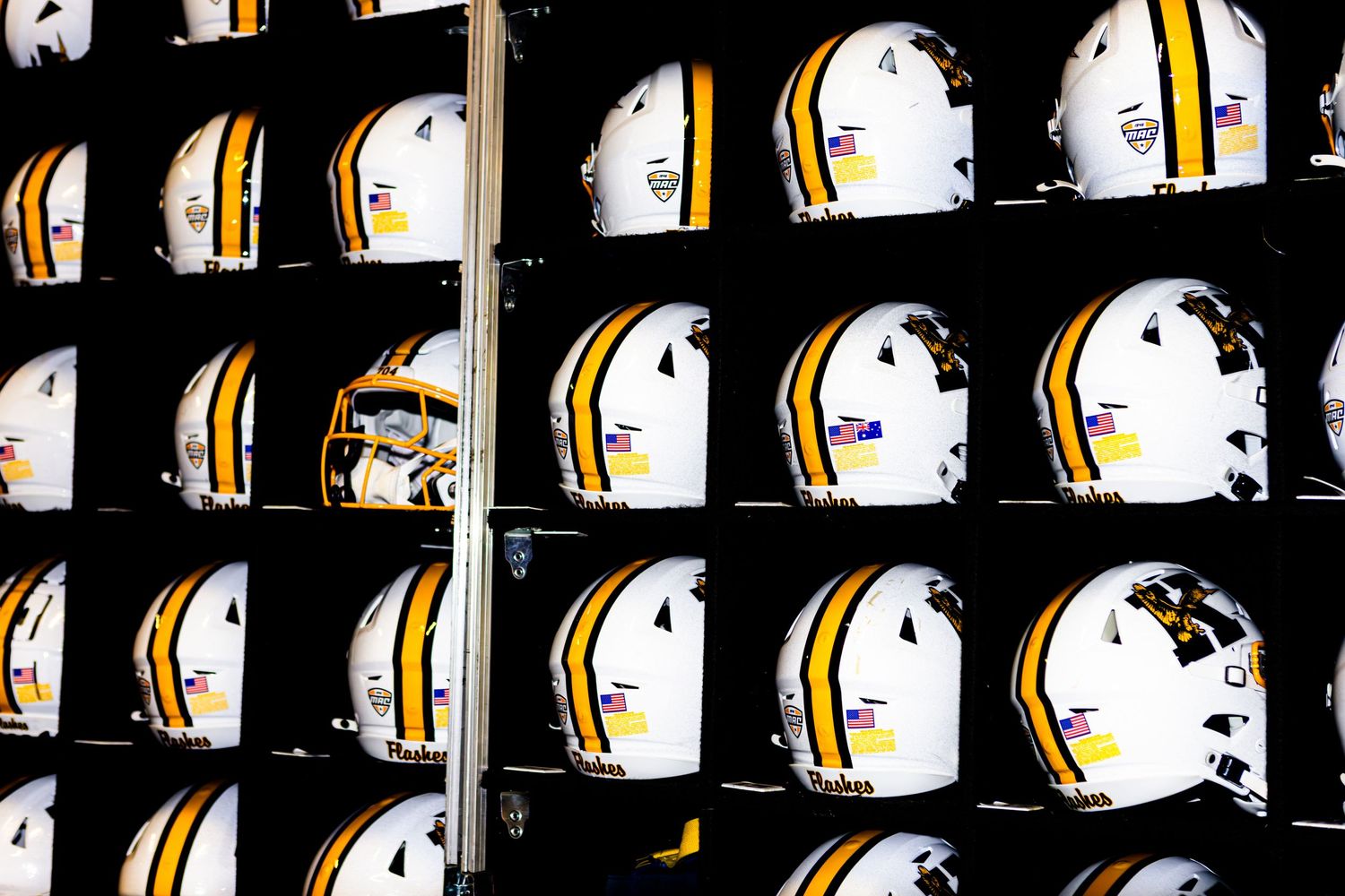 Rows of white football helmets with black and orange stripes line up on metal shelving in a sports equipment room.