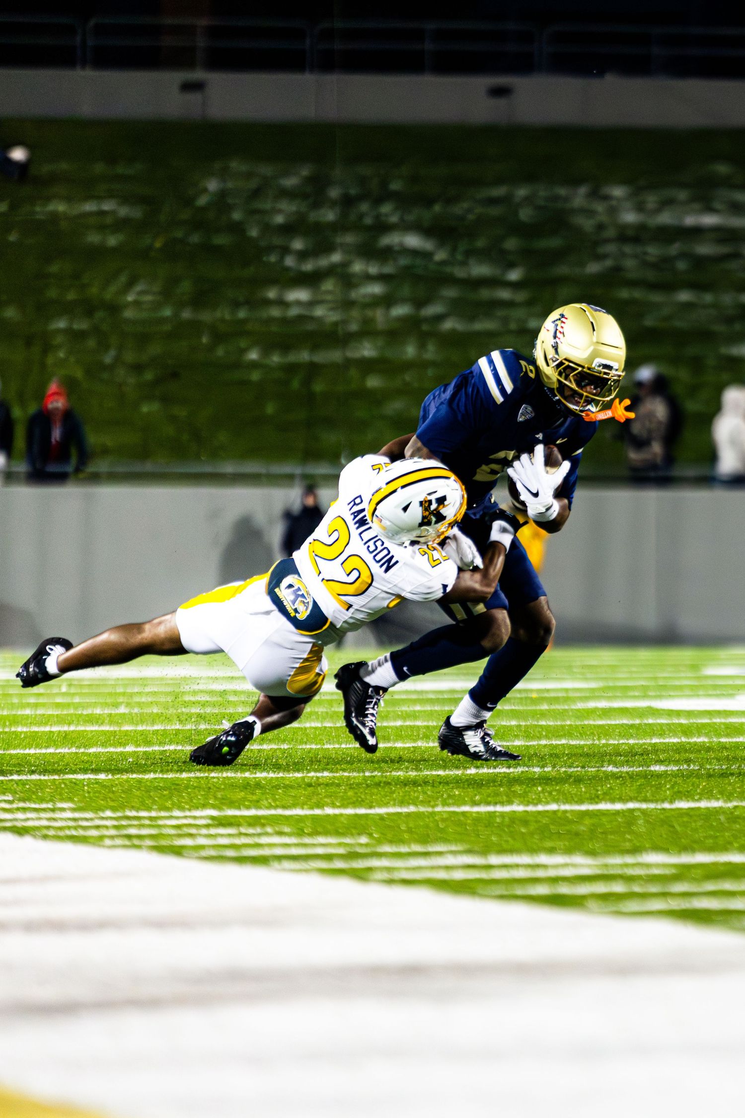 Football player in navy and gold uniform makes diving tackle attempt on field during competitive game action.