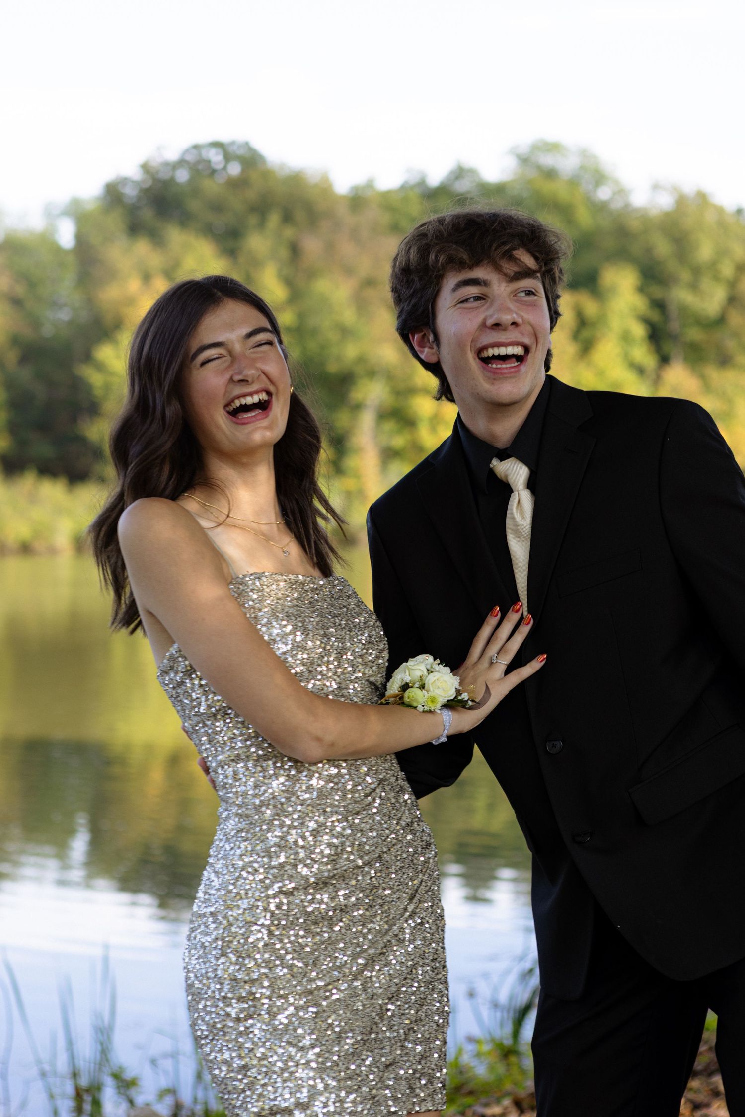 A couple in formal attire shares a candid laughing moment by a lake during a photo session.