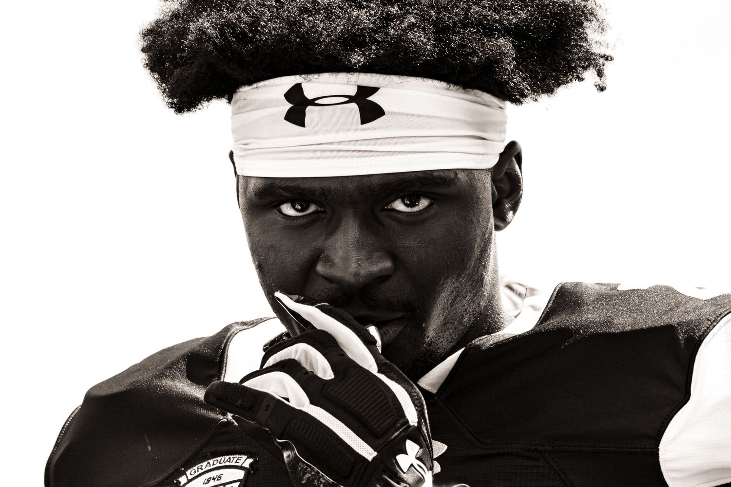 A dramatic black and white sports portrait shows an athlete wearing an Under Armour hat shot in profile against dark shadows.