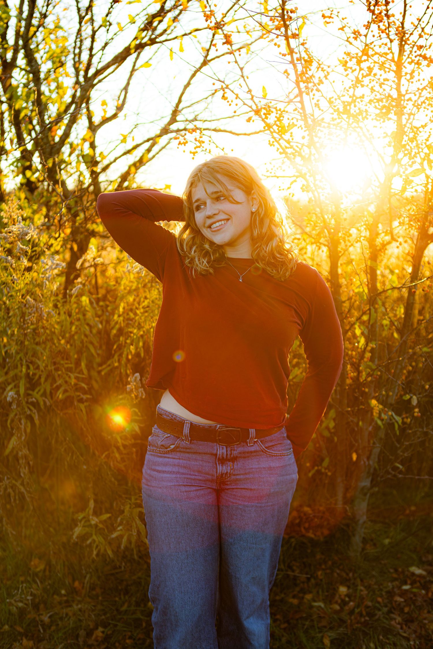 Person in a red sweater and jeans poses outdoors during golden hour sunset with sunlight streaming through autumn trees.