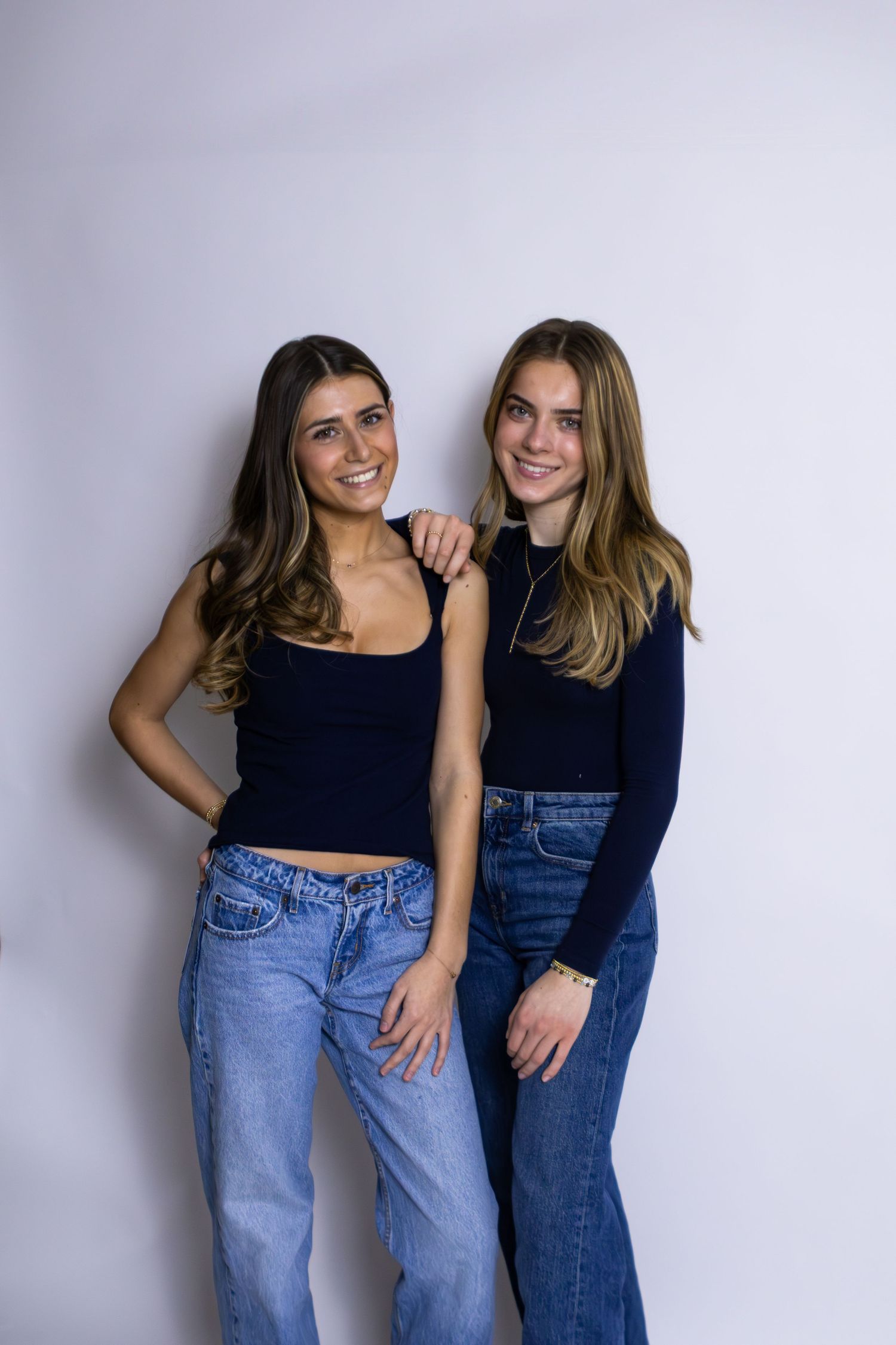 Two young women wearing jeans and casual tops pose together against a light gray background in a studio setting.