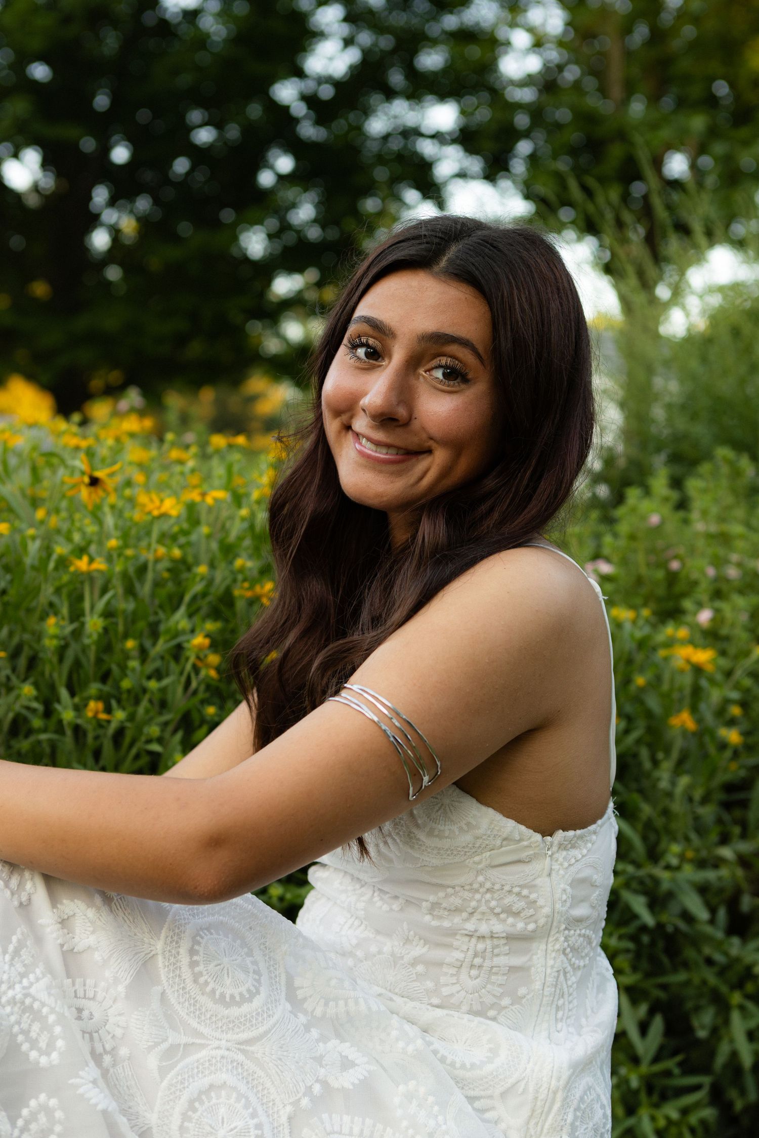 A smiling person in a white lace dress poses in a garden with yellow wildflowers and greenery in the background.