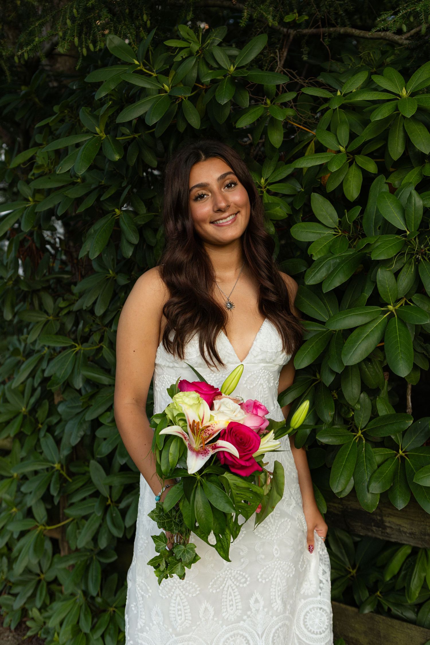 A person in a white lace wedding dress holds a vibrant bouquet with pink lilies against a backdrop of green foliage.