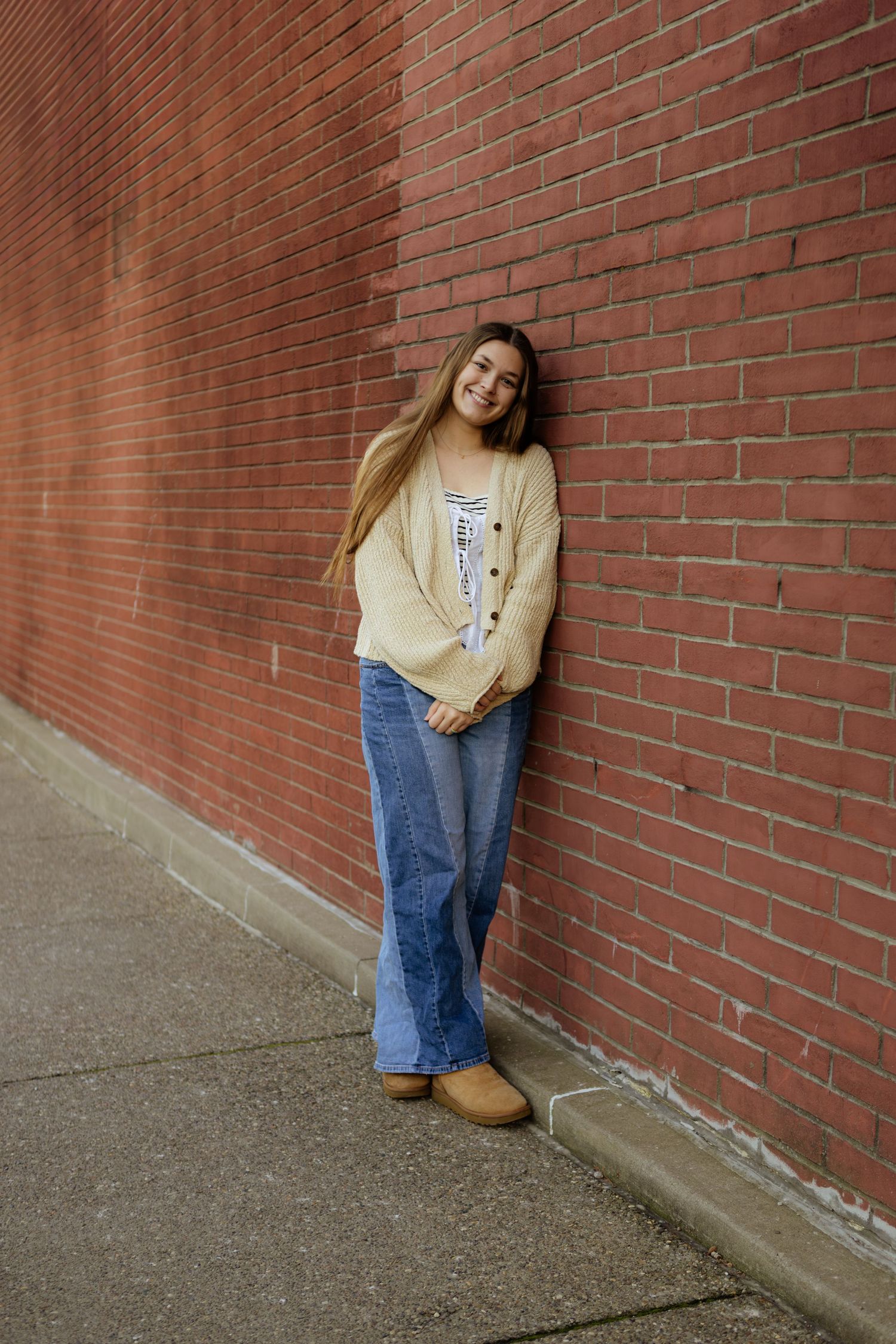 A person in flared jeans and a beige sweater leans against a red brick wall on a city sidewalk.