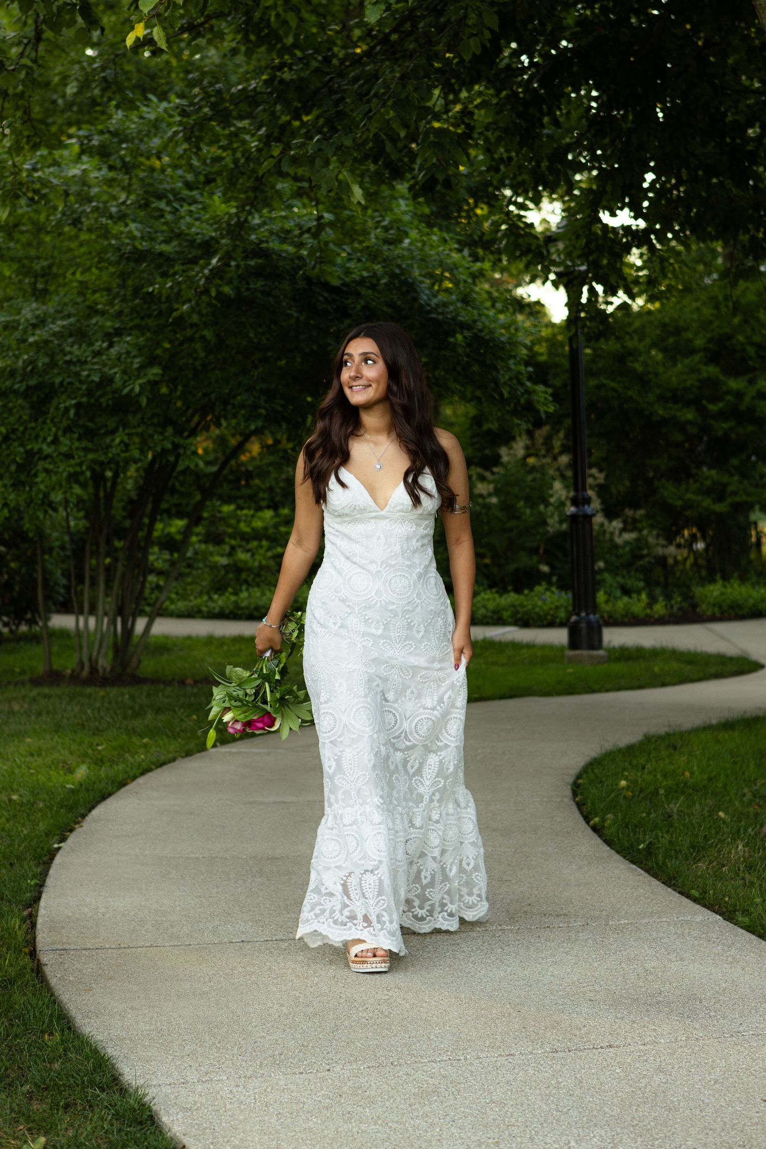A person in a white lace dress poses gracefully on a curved garden path surrounded by greenery and flowers.