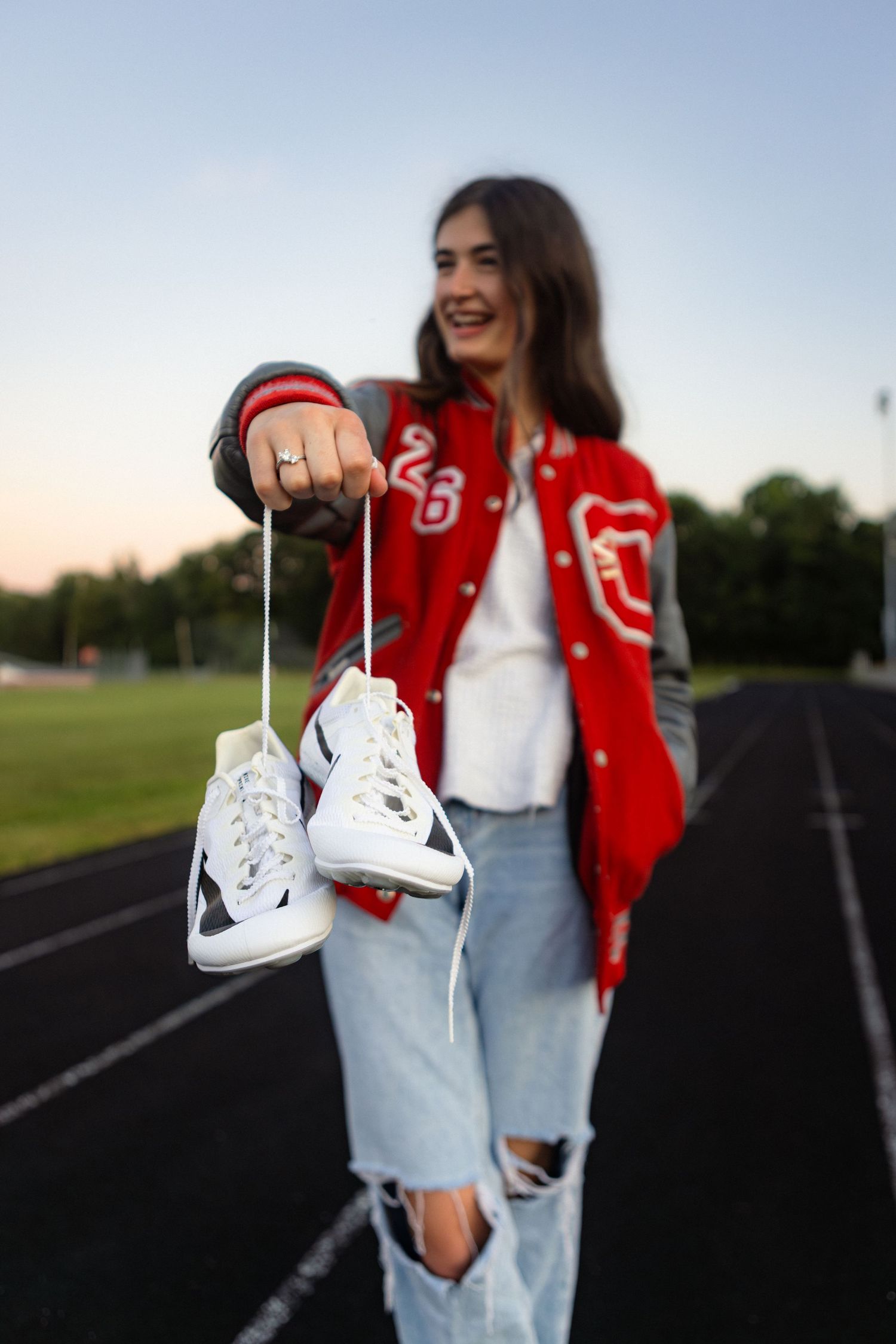 Someone in a red varsity jacket and ripped jeans holds up a pair of white sneakers on an outdoor running track at sunset.