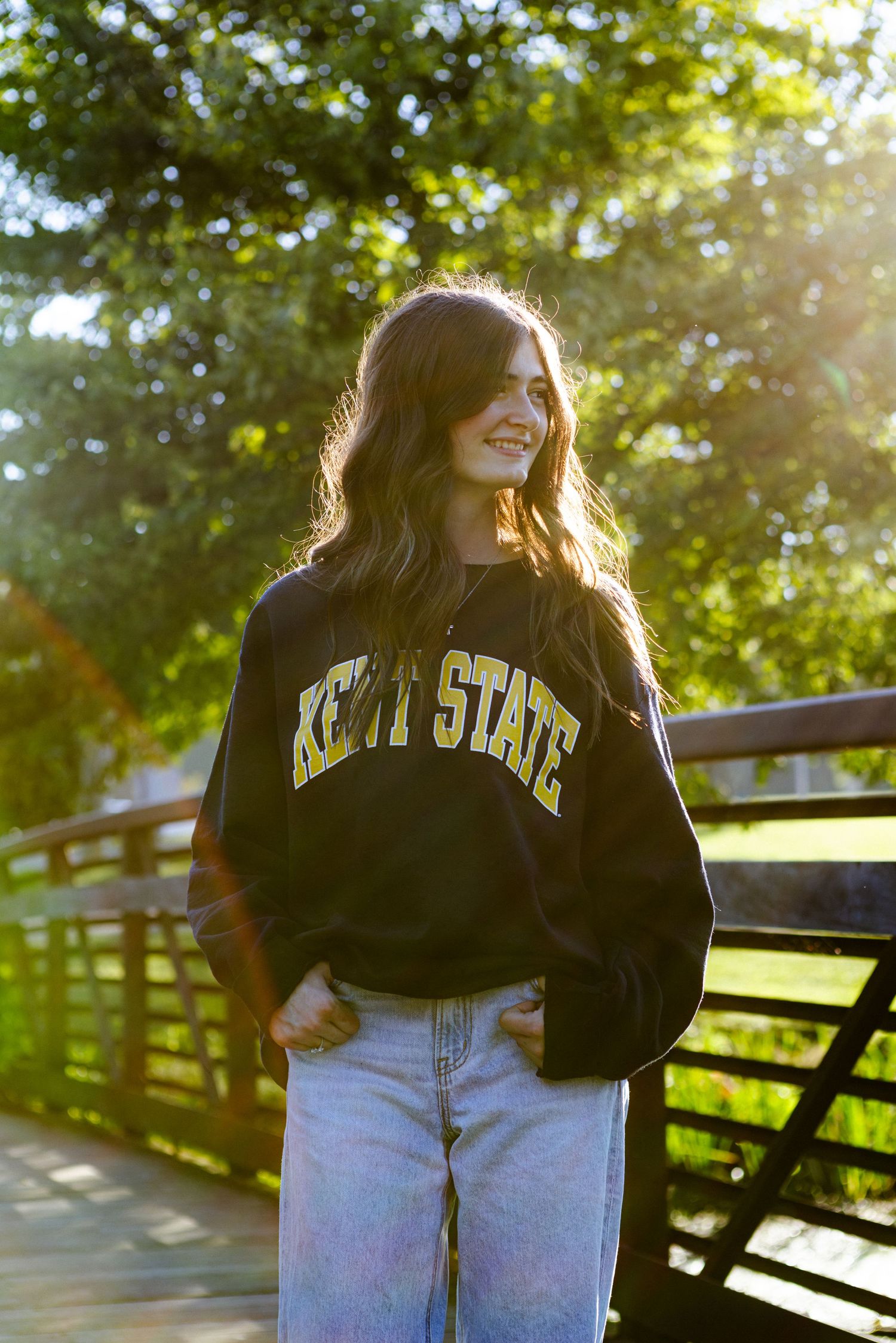 Casual college student in Kent State sweatshirt and jeans leaning against wooden railing in sunlit outdoor setting.