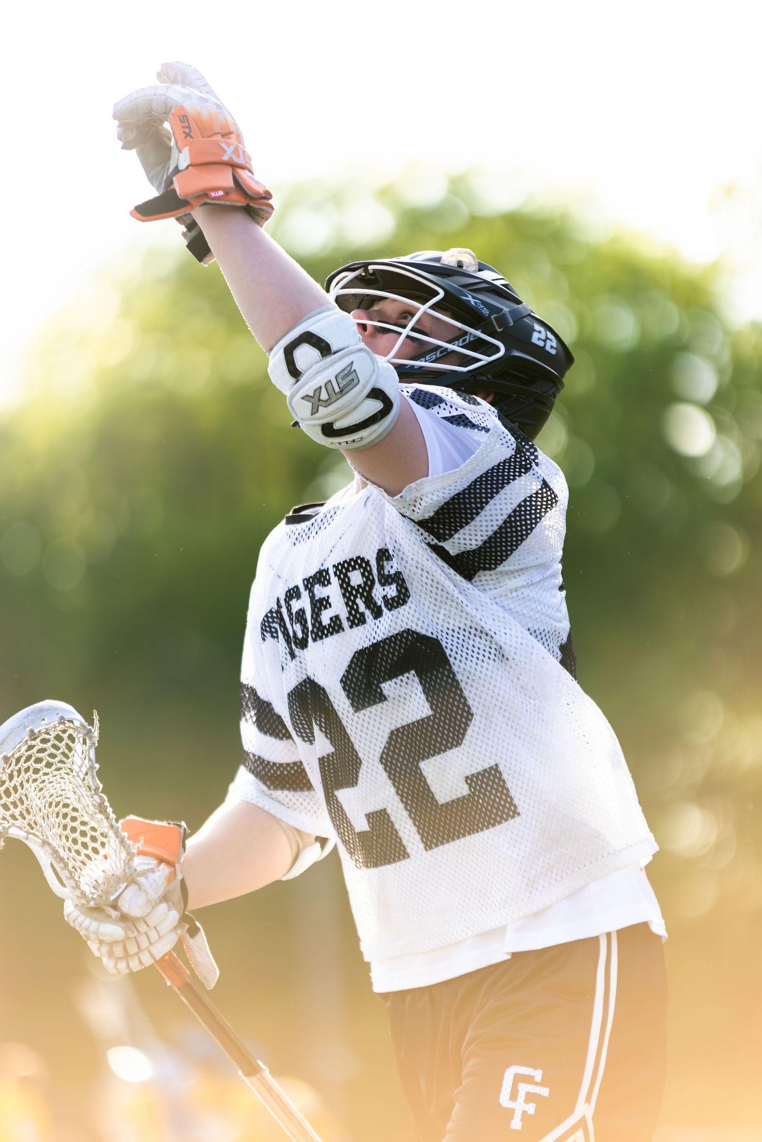 Lacrosse player in white jersey celebrates with raised stick at sunset.