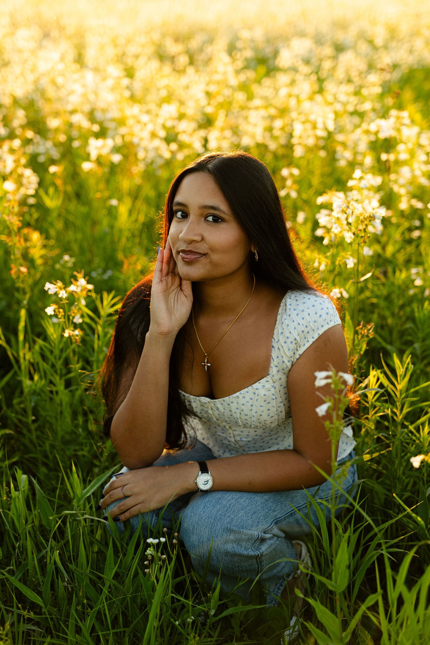 Young woman in white floral top and jeans crouching in a sunlit wildflower field, posing thoughtfully.