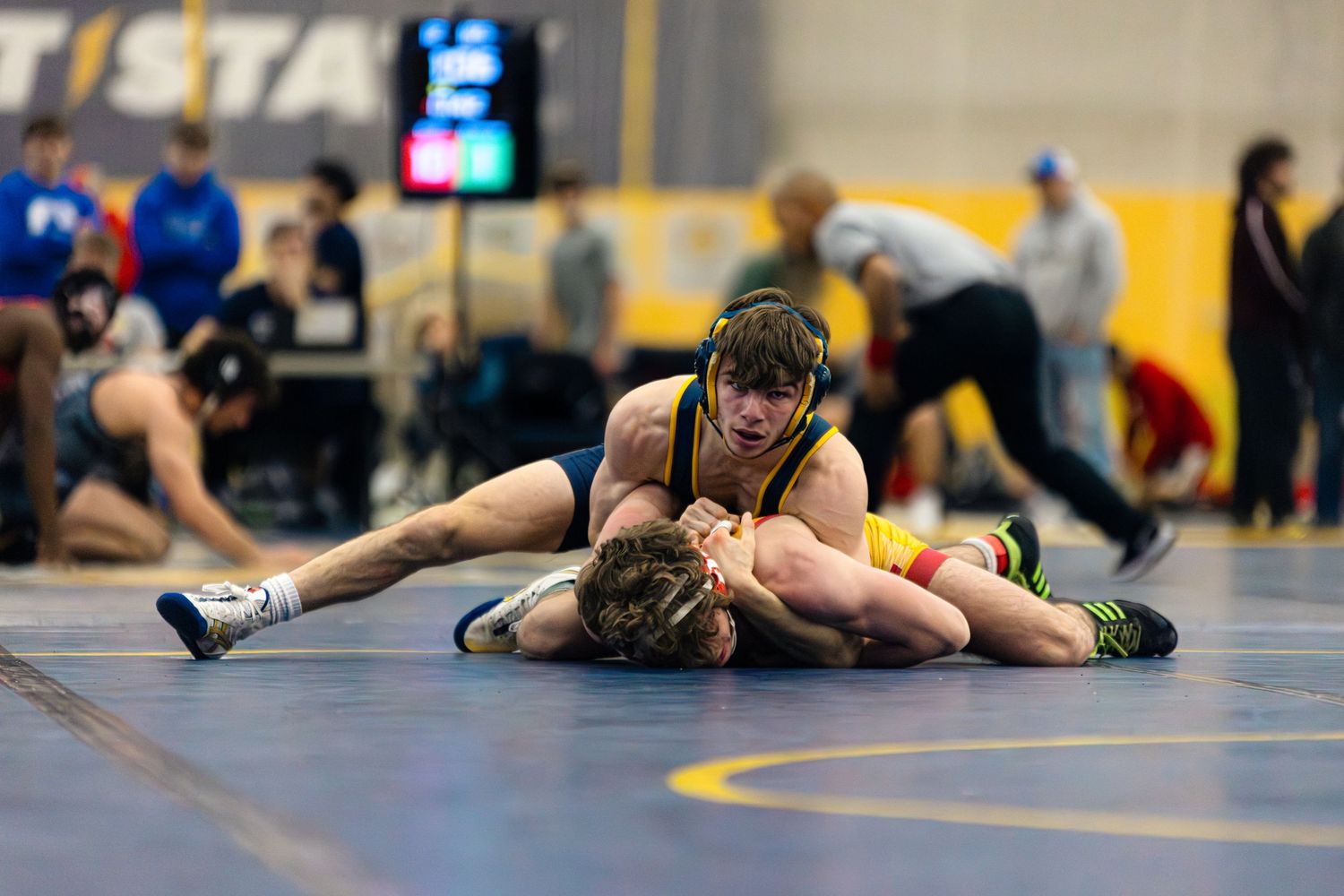 Two wrestlers grappling on a blue mat during a competitive match at an indoor sports venue.