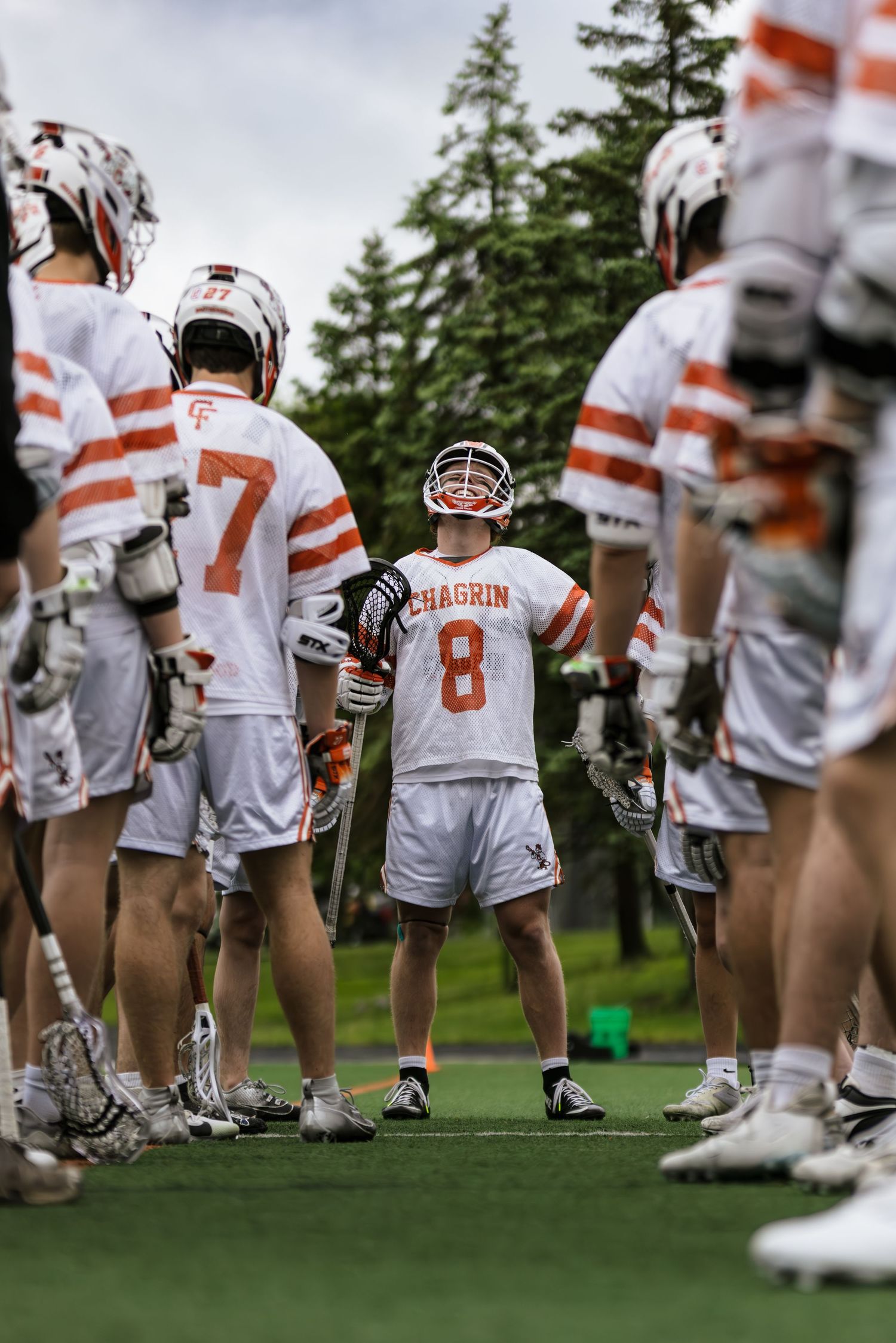 College lacrosse team players in orange and white striped jerseys line up on a grass field during practice.