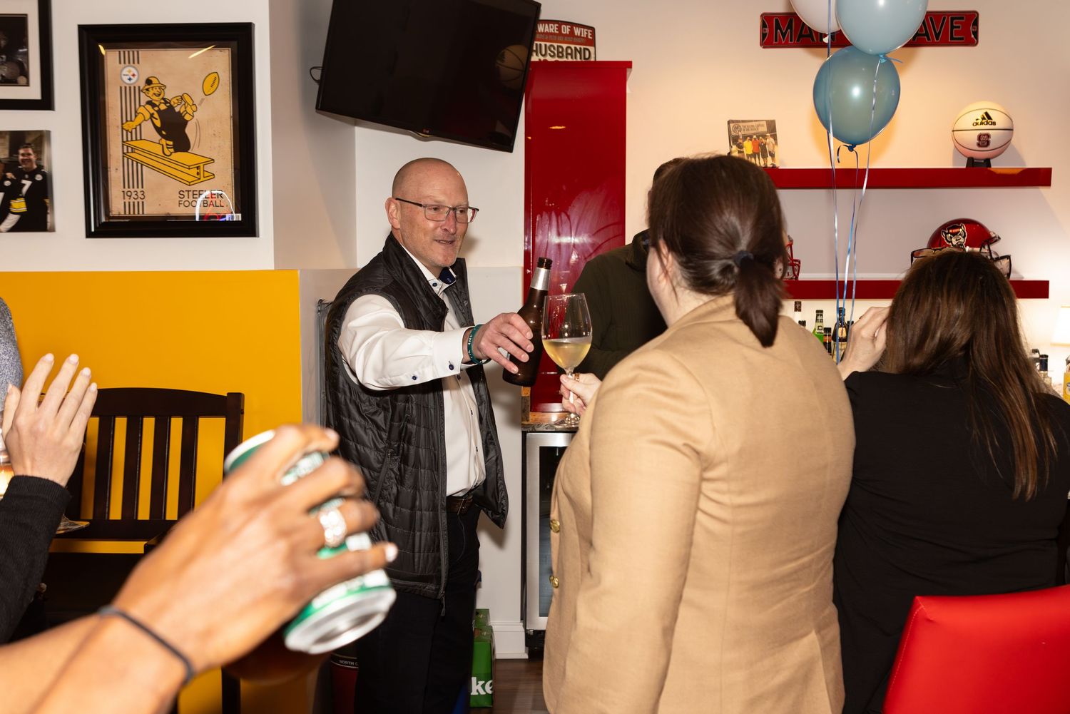 A group gathered at a social event with blue balloons visible while someone gestures with a drink in hand.