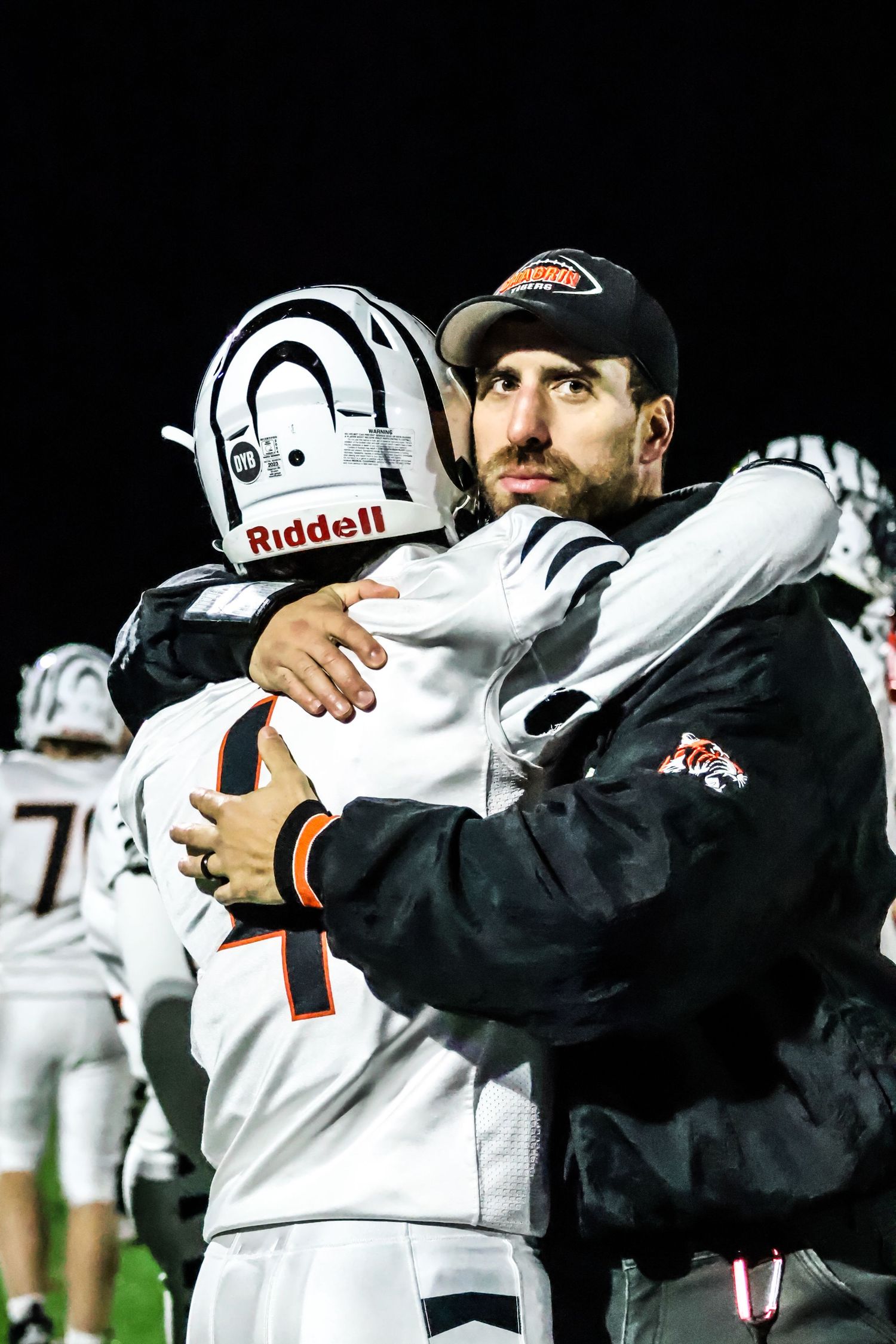 Football coaches in black jackets and white helmets huddle on the sidelines during a night game.