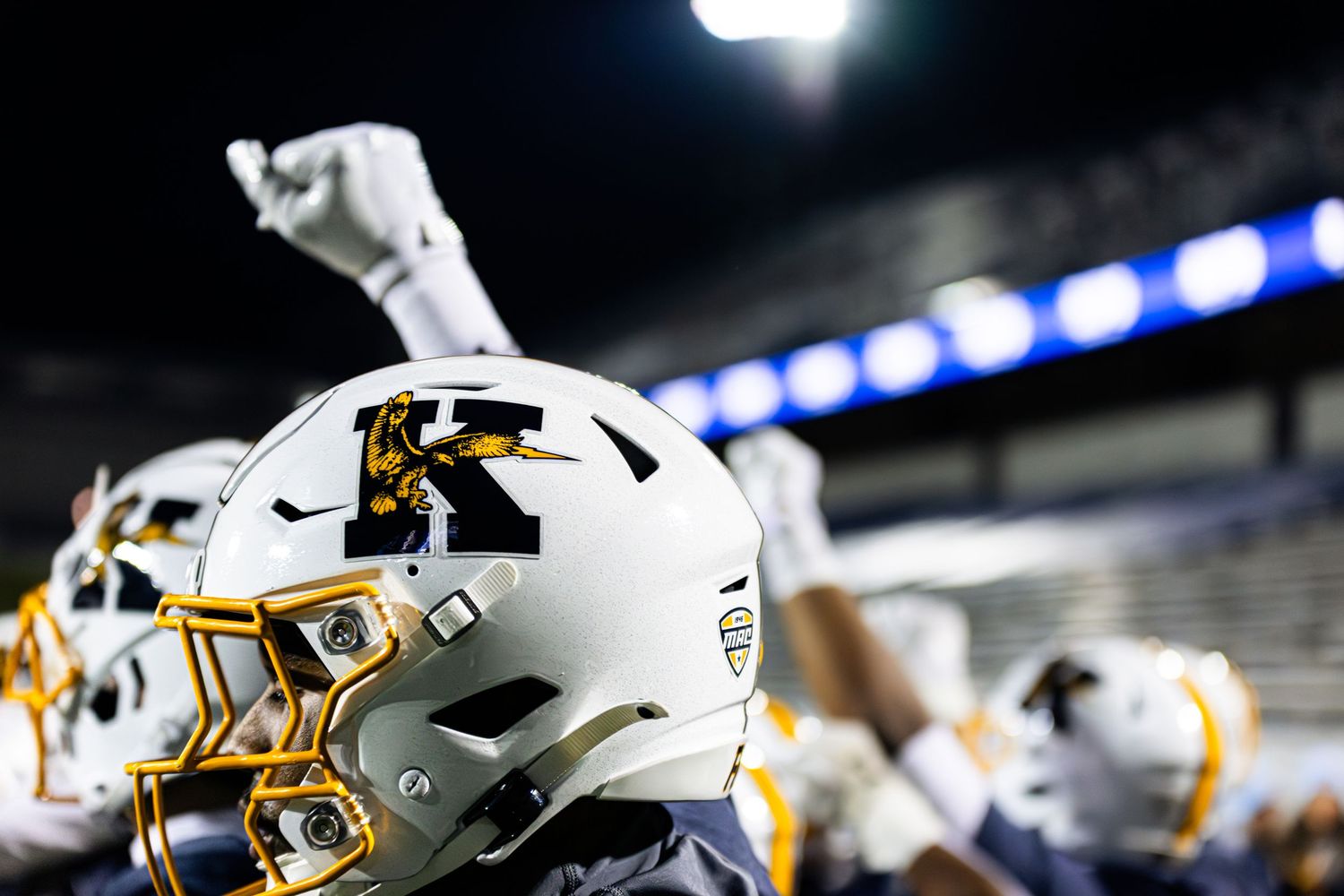 A series of football players in white and gold uniforms lined up on the field during a night game.