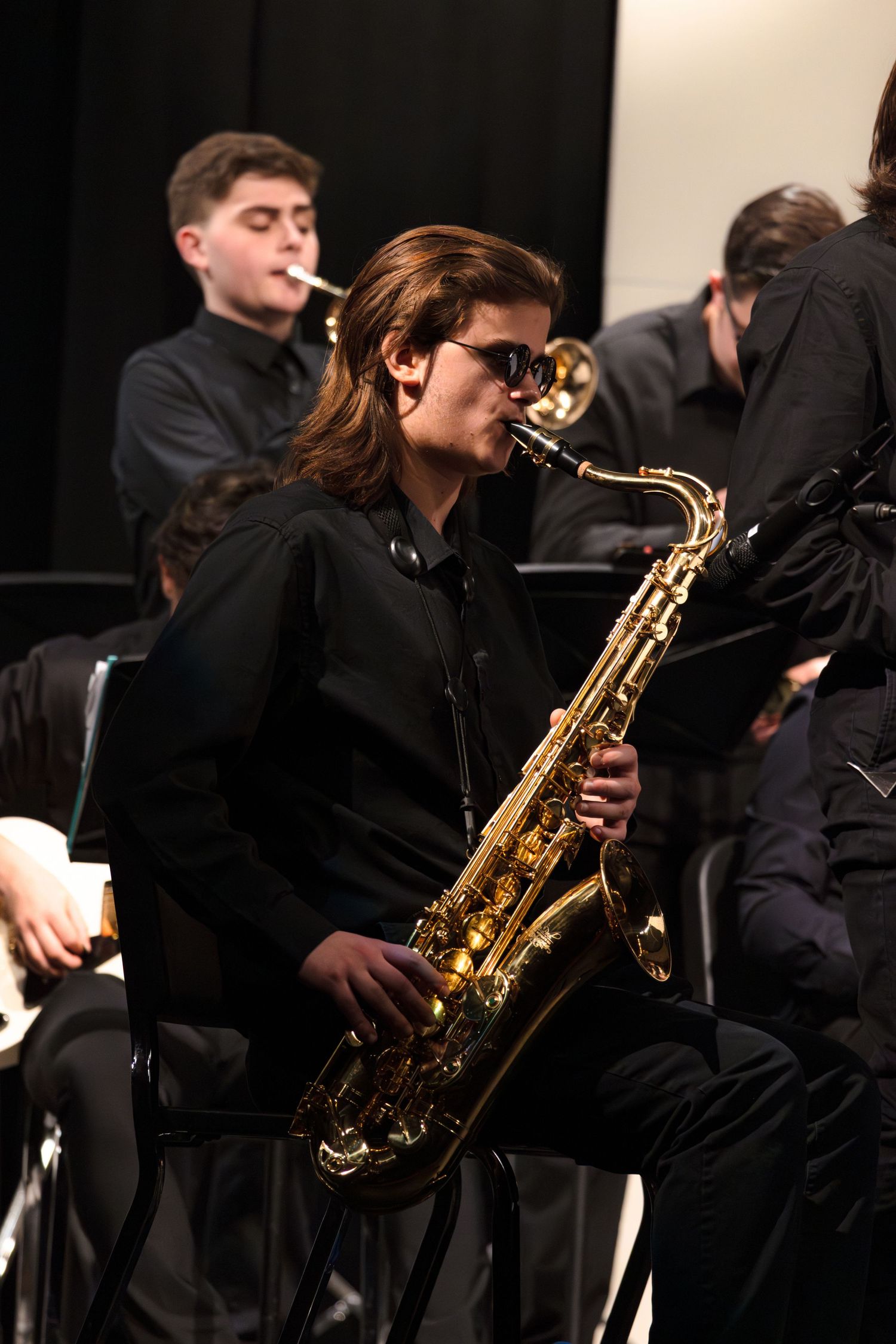 Student musicians perform with saxophones during a jazz band concert while wearing formal black attire.
