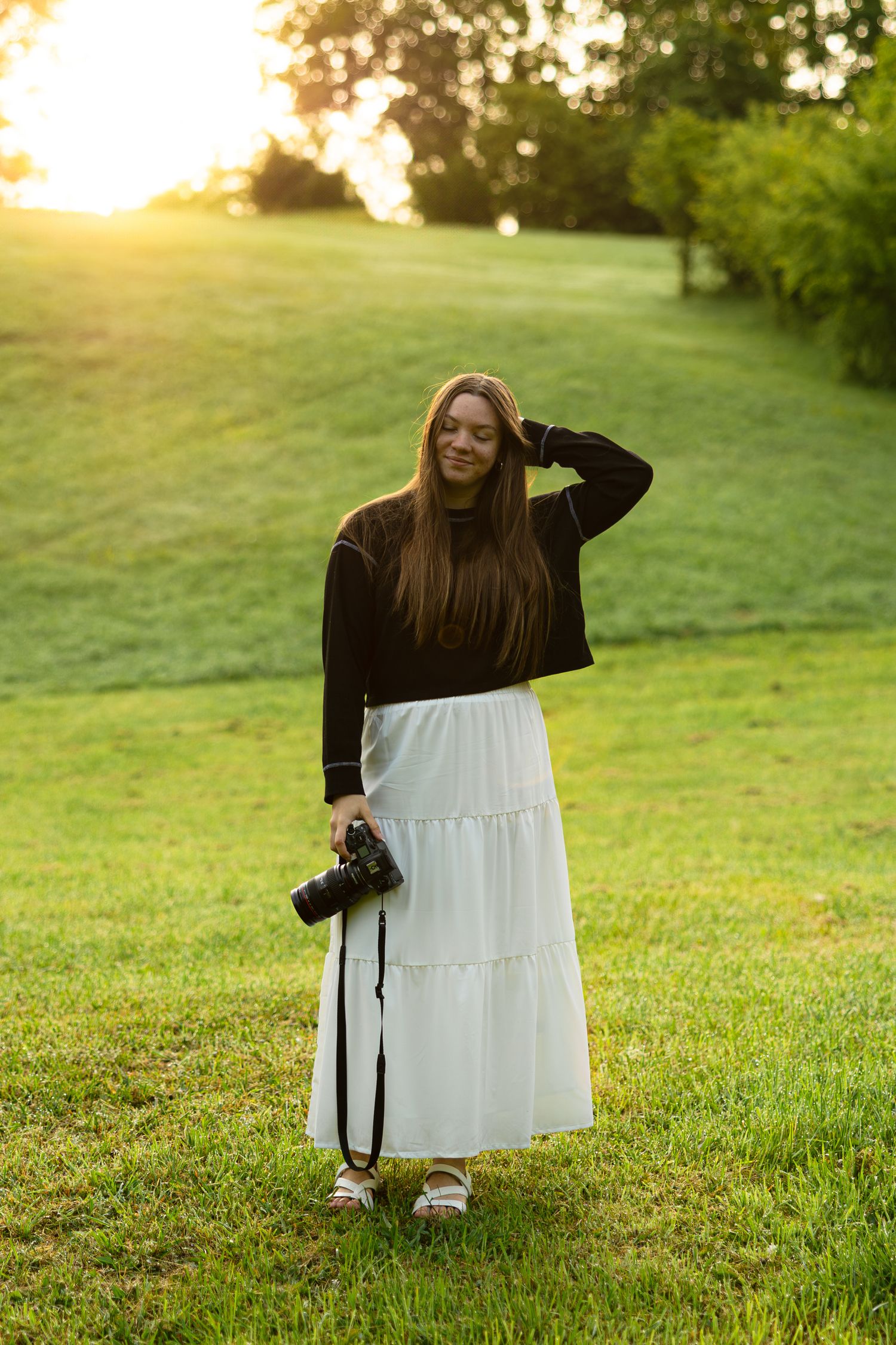 Photographer in black sweater and white skirt stands in a sunlit meadow with camera equipment.