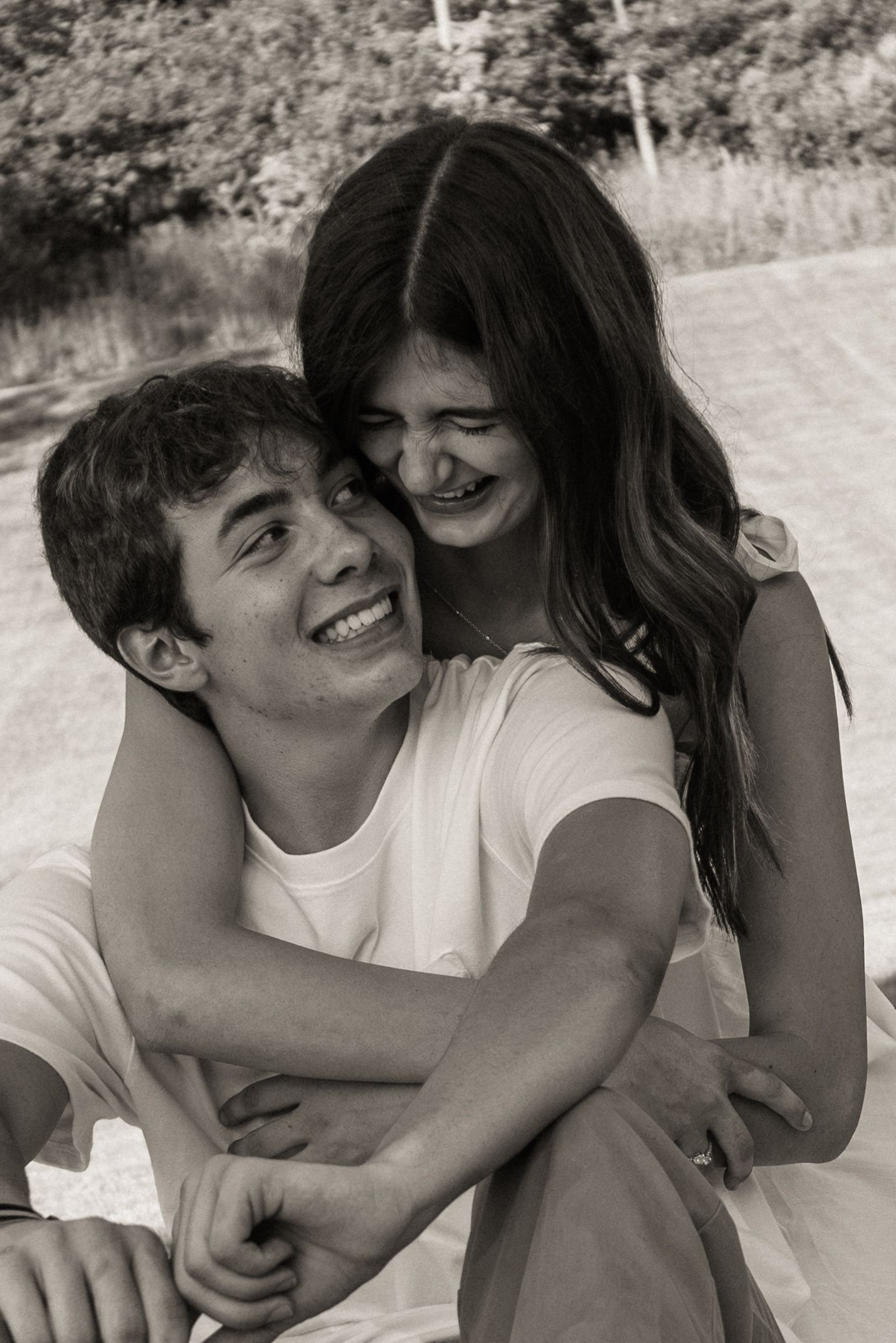 A candid black and white photo shows a playful young couple embracing and laughing together outdoors.