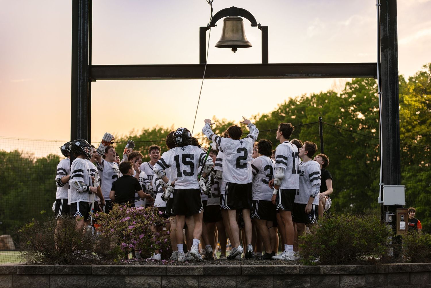 A group of athletes gathered around a victory bell at sunset.
