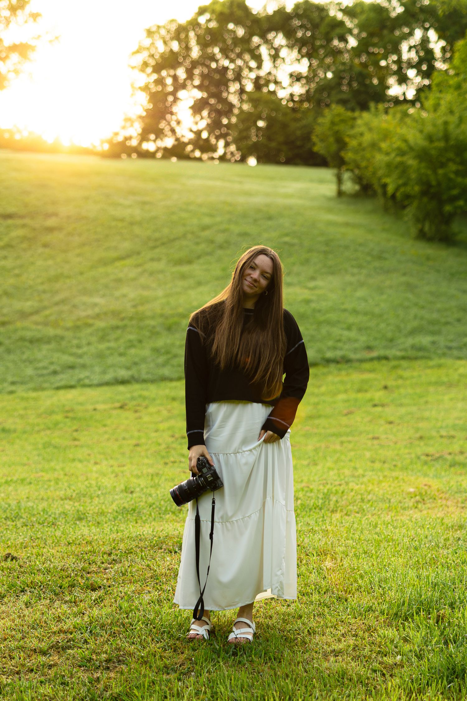 Someone in a white dress and black boots crouches in a grassy field at sunset while creating various poses.