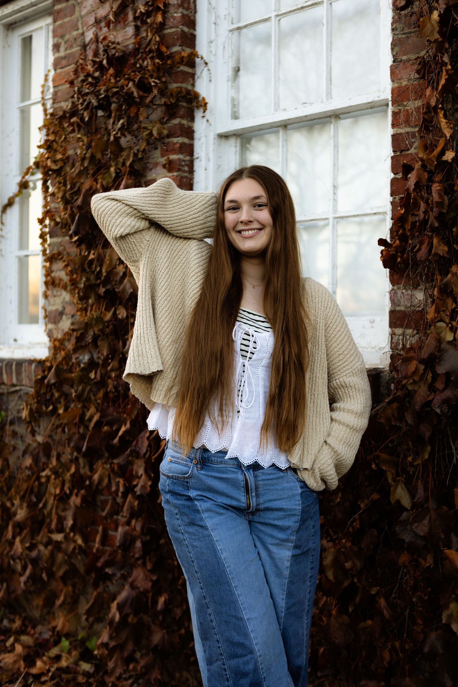 A person in a beige sweater and jeans poses playfully against a brick wall with autumn ivy.