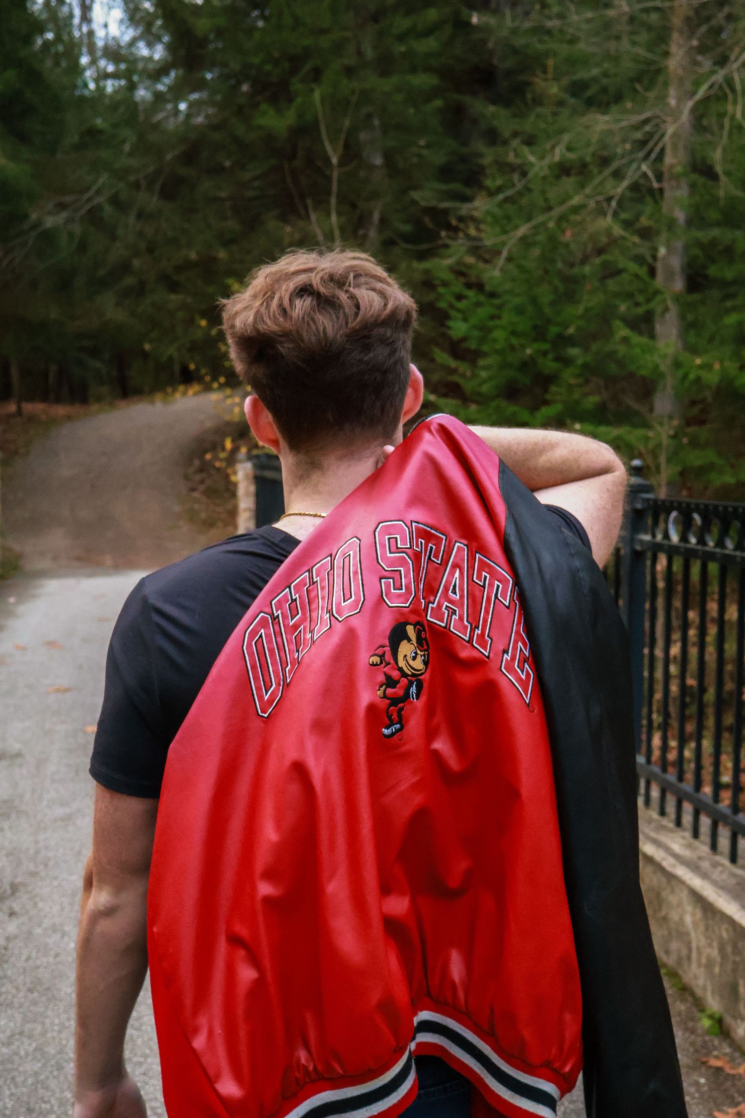 Person wearing an Ohio State red varsity jacket walks along a wooded path on a cloudy day.