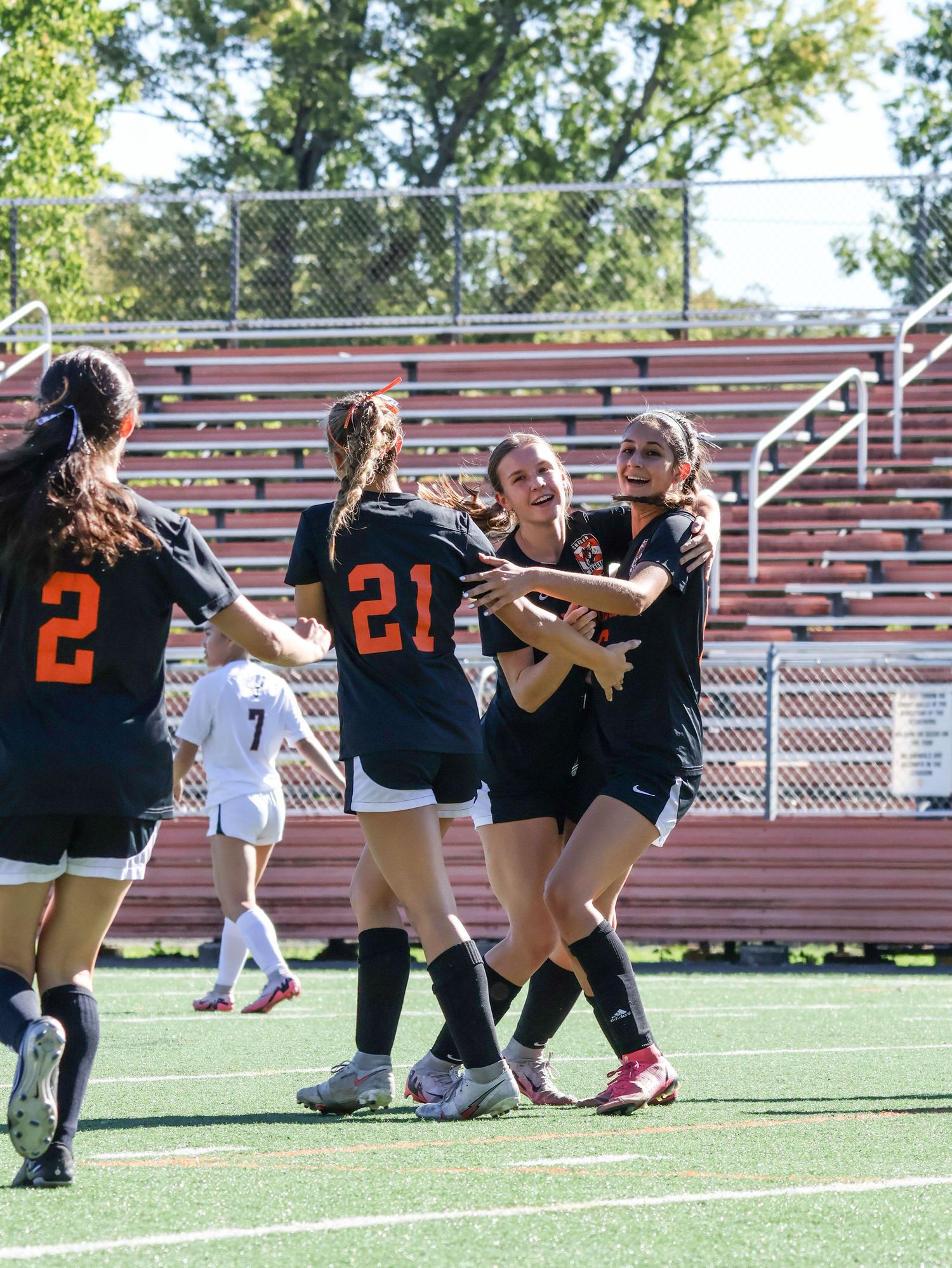 Dancers in black jerseys perform synchronized kicks and movements on a sports field with red bleachers in the background.