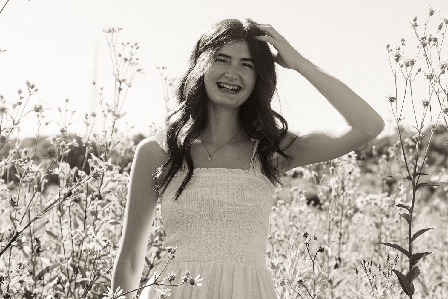Black and white portrait of someone smiling joyfully while standing in a field of wildflowers at sunset.