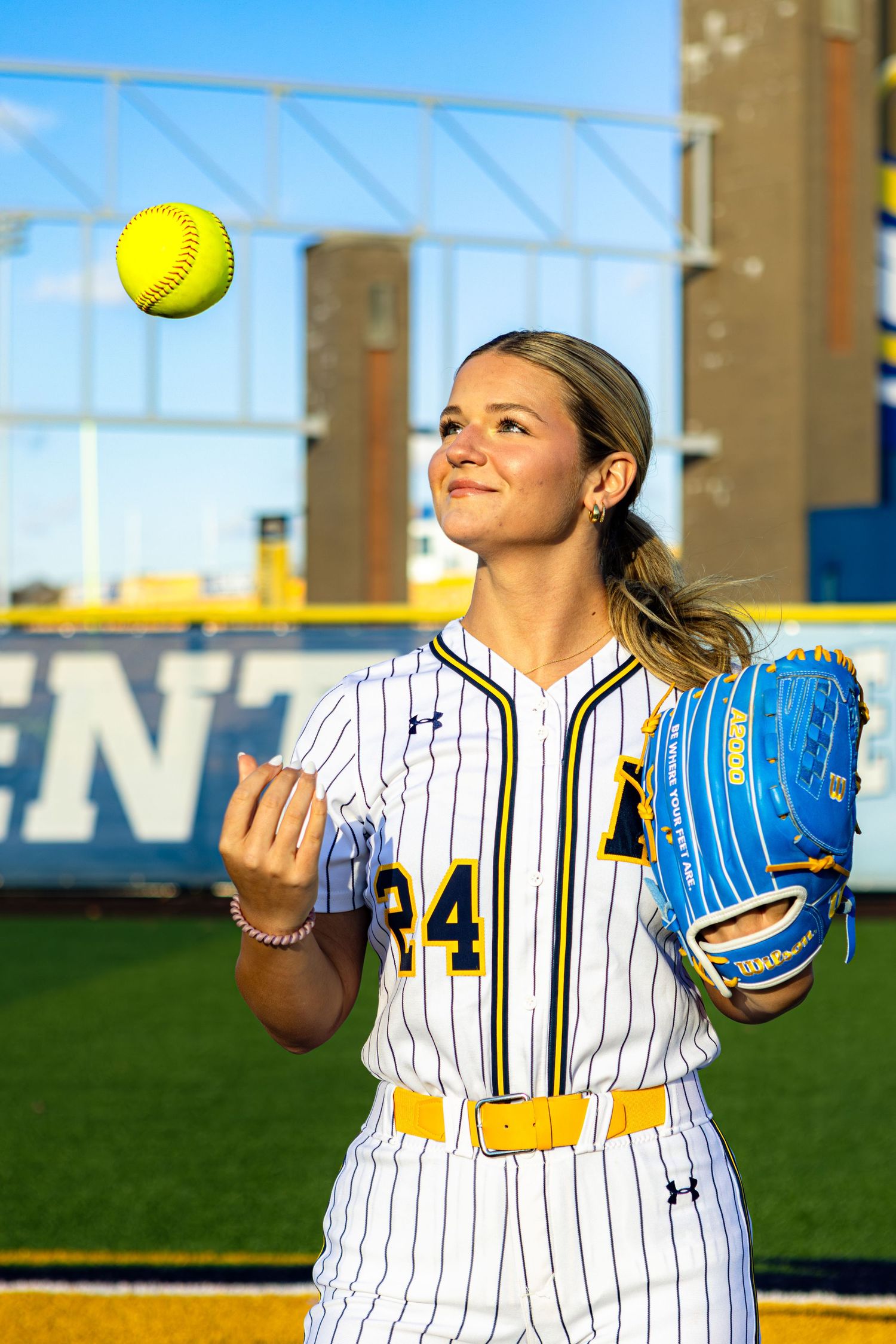 Softball player in pinstriped uniform demonstrates pitching sequence with yellow ball against blue sky background.