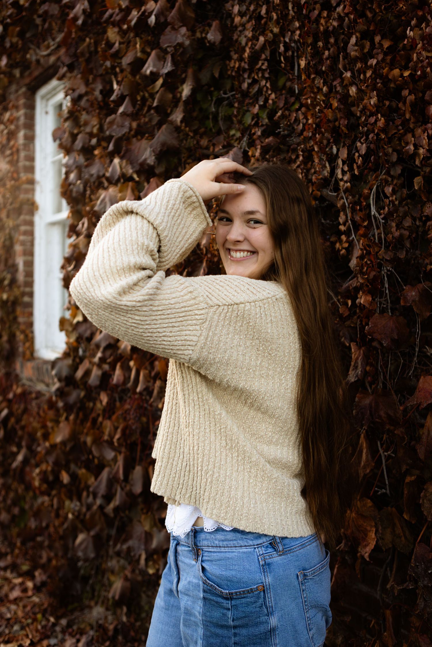 Person in cream knit sweater and blue jeans poses playfully against autumn ivy covered wall.