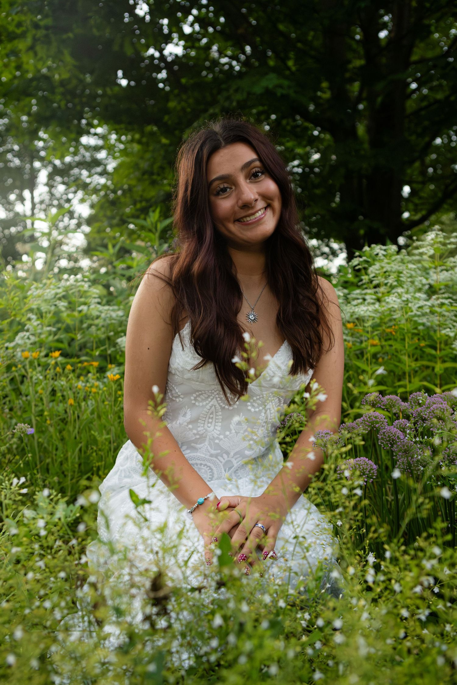Someone wearing a beautiful white lace wedding dress poses in a garden meadow surrounded by wildflowers and greenery.