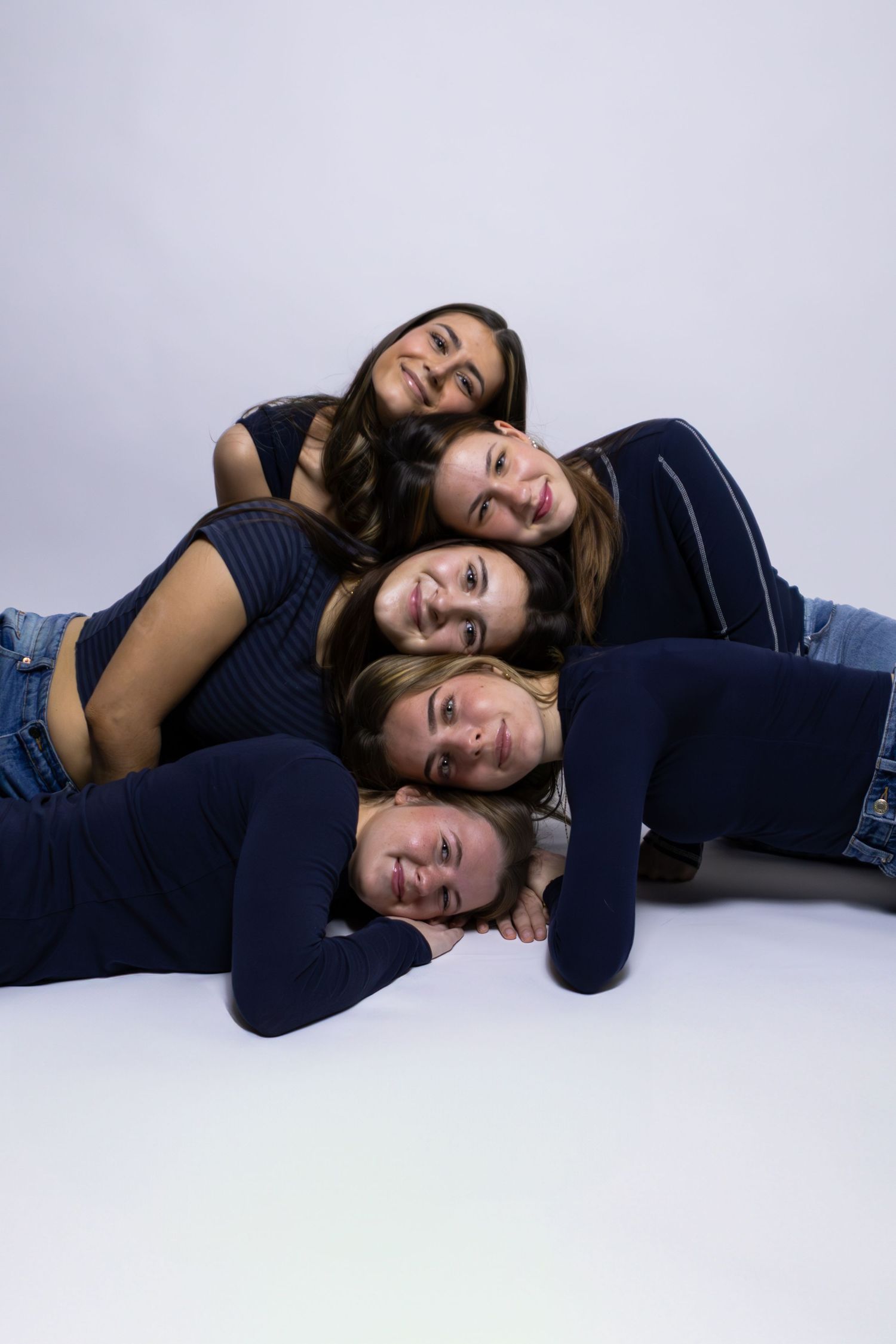 A group of people in navy blue sweaters lying down in a playful stack pose against a light gray background.