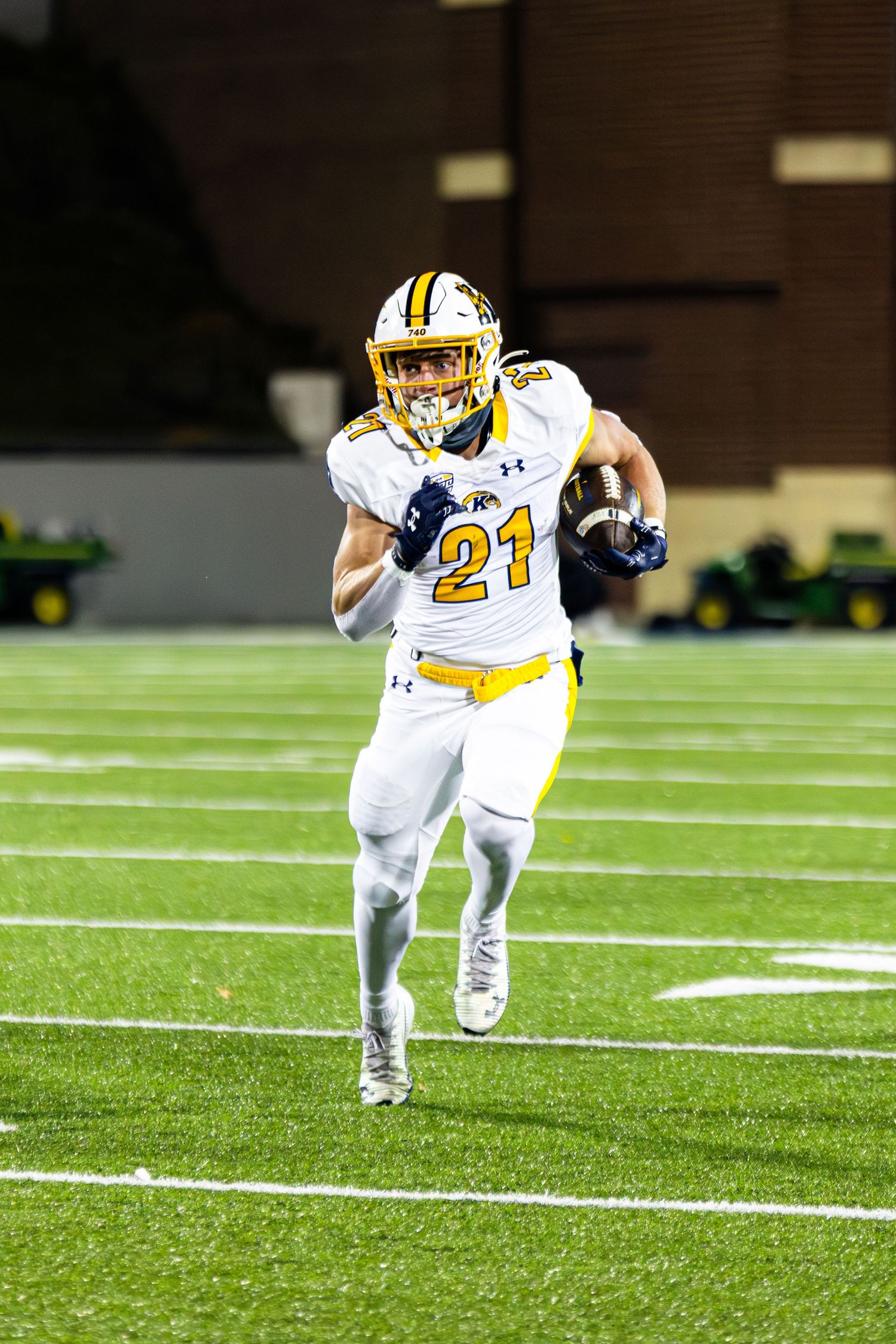 Football player in white and yellow uniform number 21 runs with the ball on artificial turf during nighttime game.