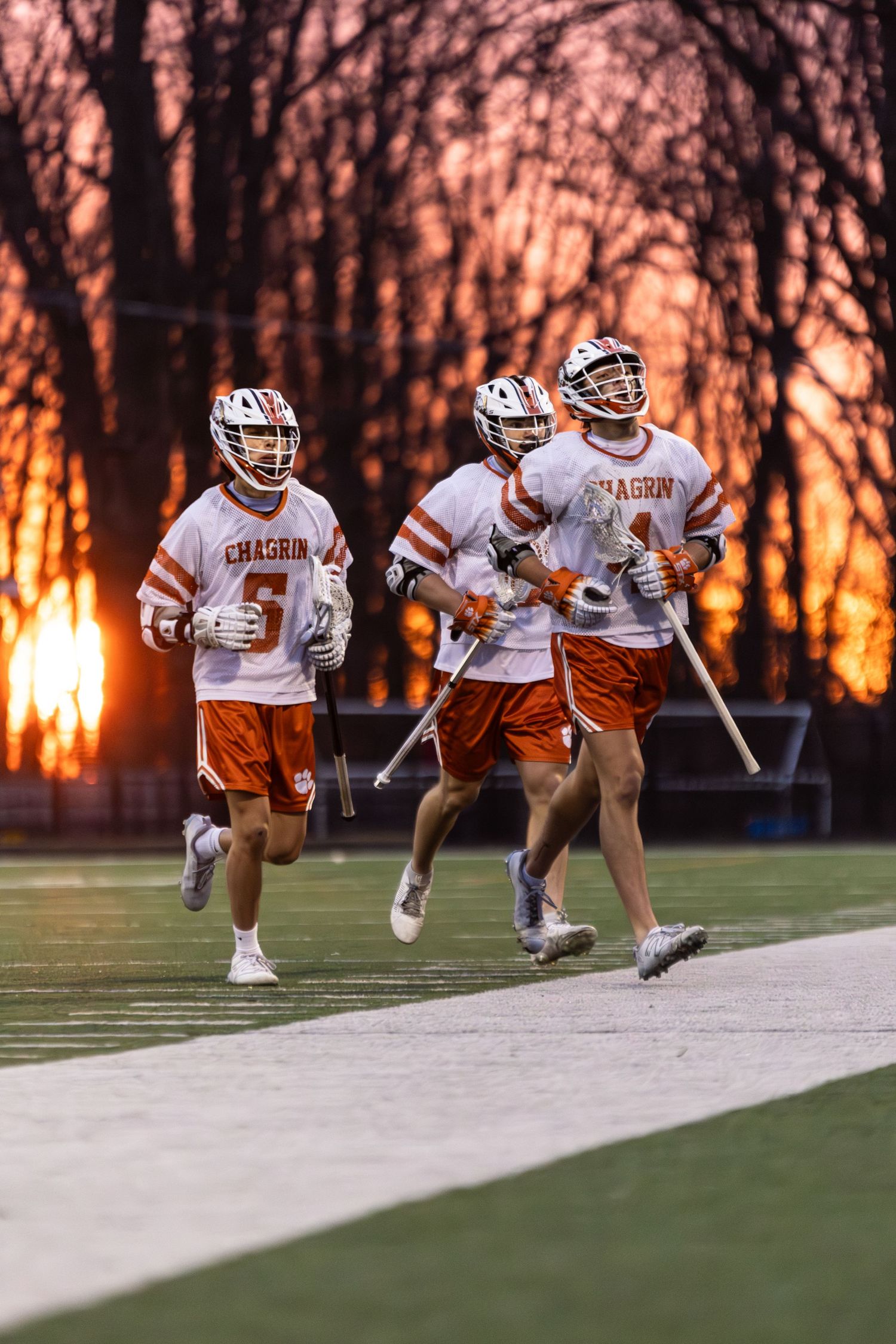 Lacrosse players running on field during sunset with dramatic orange sky.