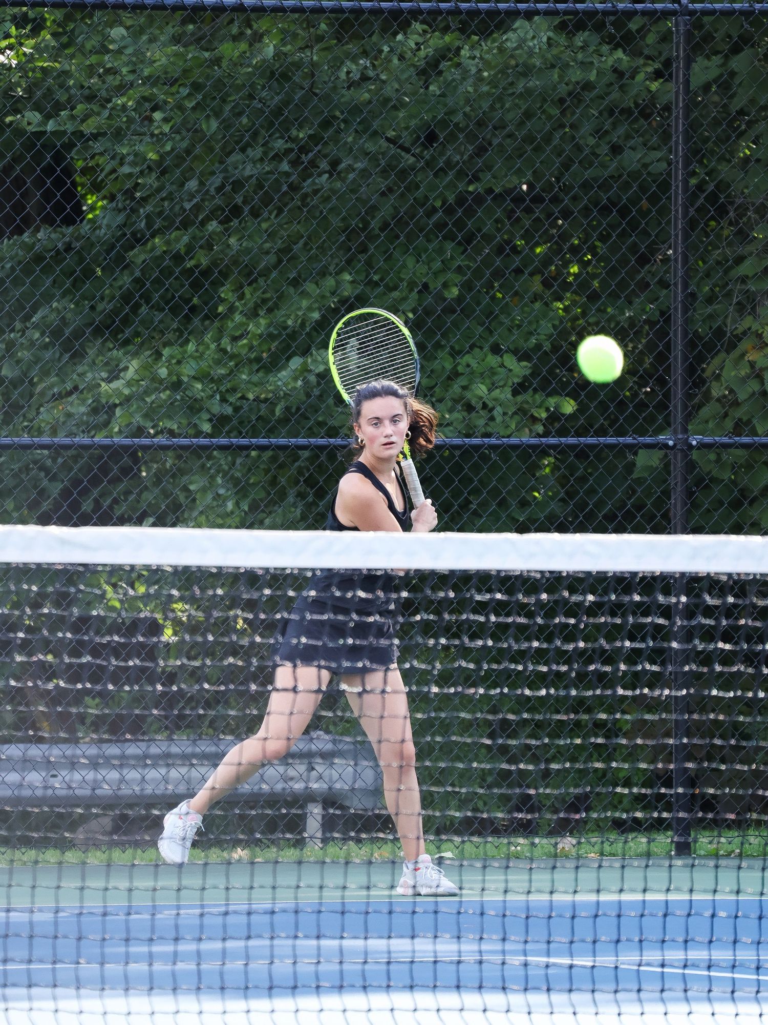 Tennis player reaches for a shot on an outdoor court with green fence backdrop.