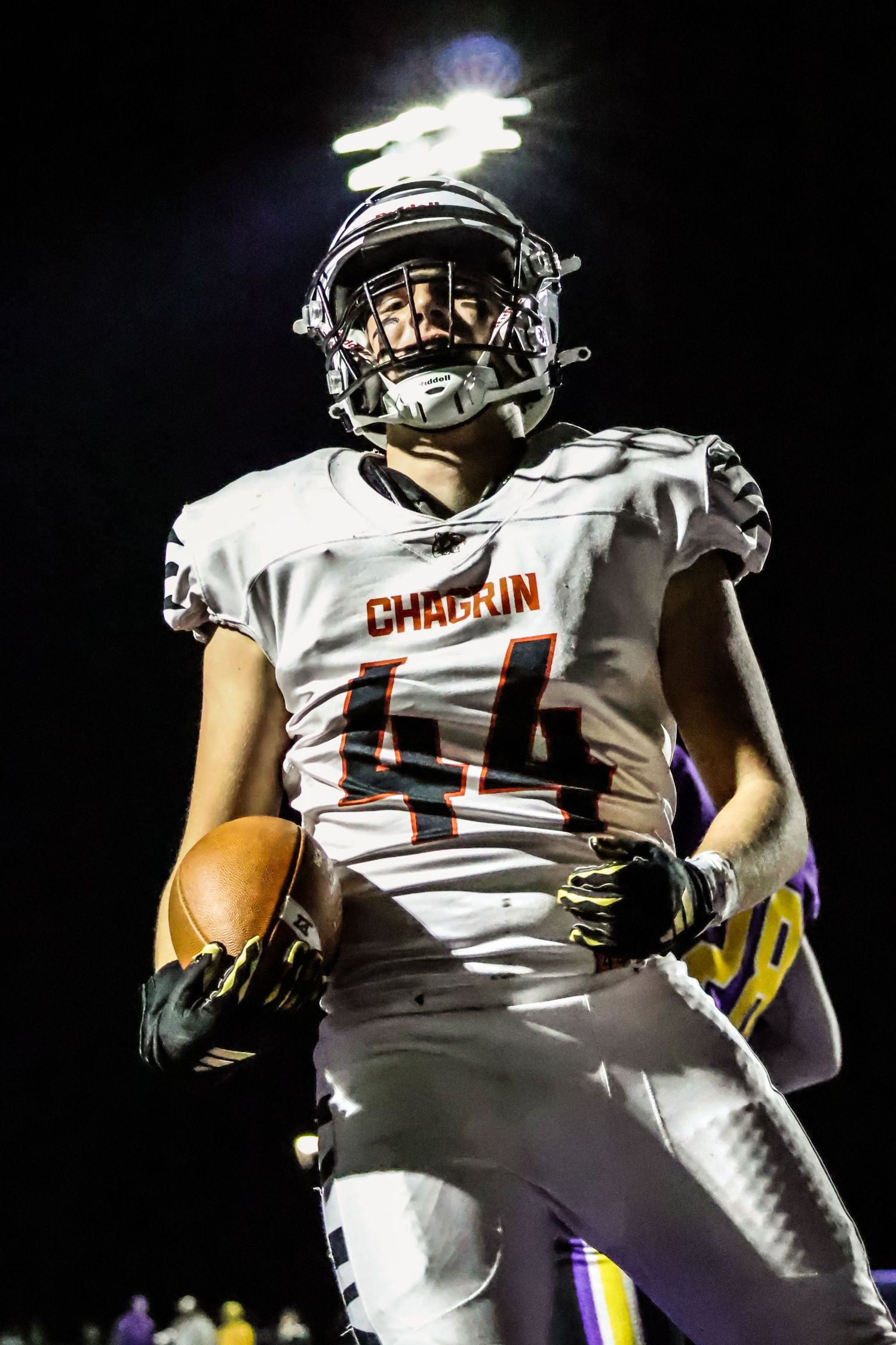 Football player in white Chardon uniform carrying ball under stadium lights.