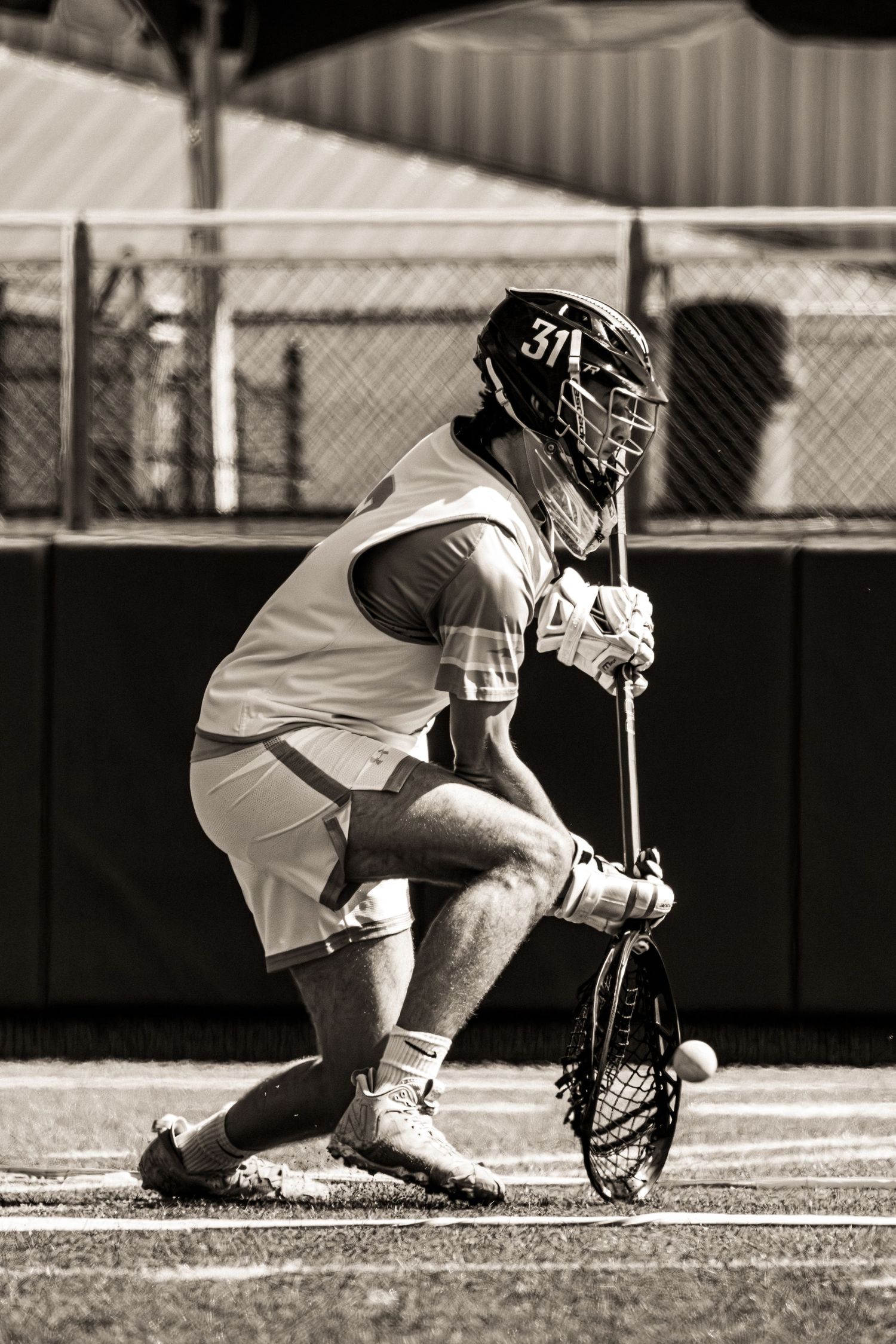 A lacrosse player in black and white uniform crouches low to scoop up the ball during a game.