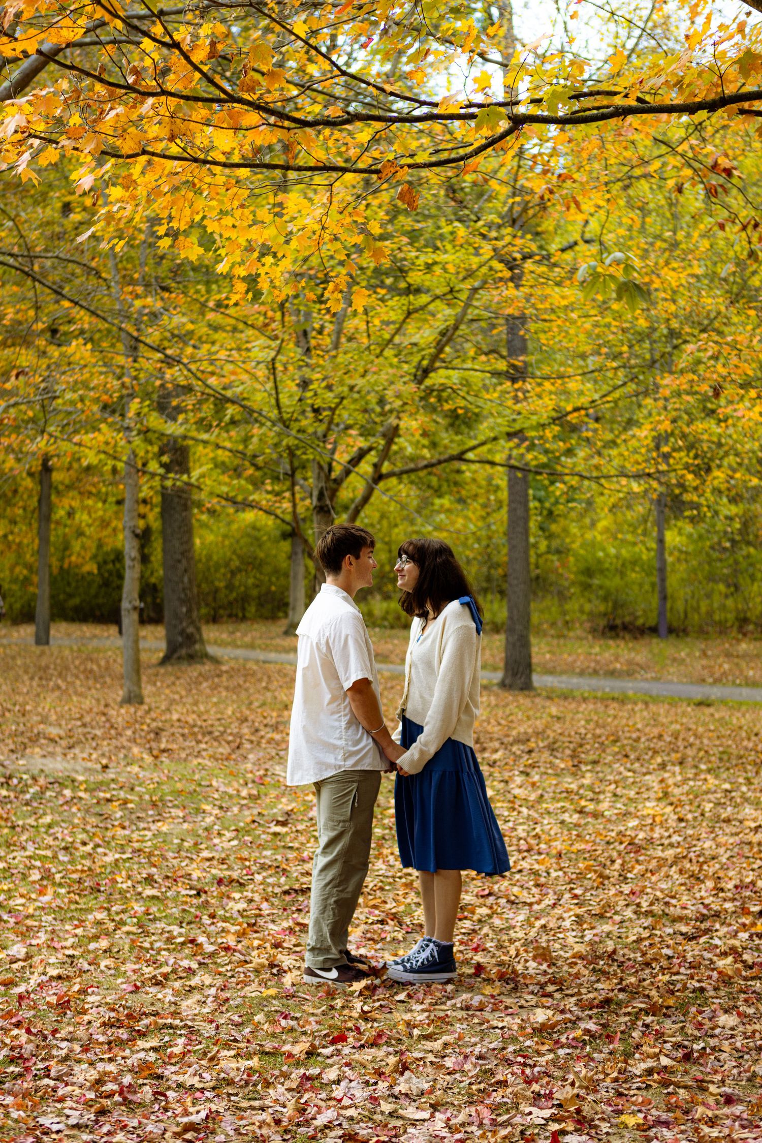 Two people stand together in a park surrounded by golden autumn leaves and trees with yellow foliage.