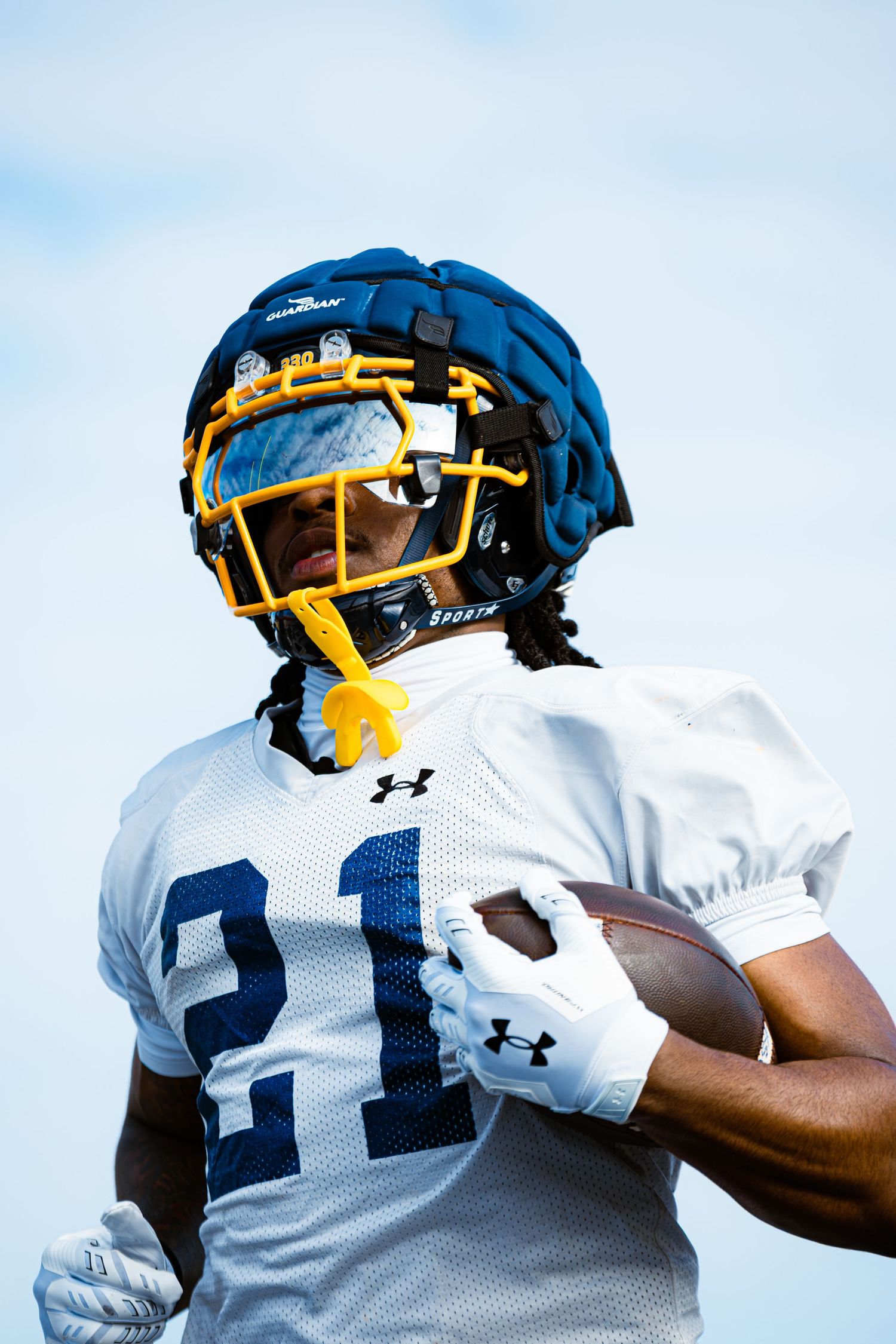 Football player #21 in white Under Armour jersey and blue helmet with yellow face mask carries ball during practice.
