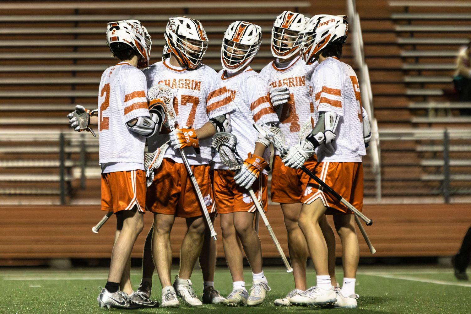 Lacrosse team huddles together on field wearing white and orange uniforms.