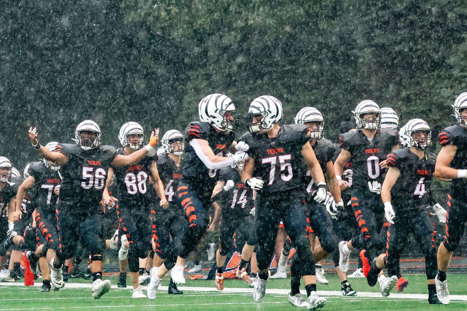 Football team in black uniforms runs together across wet field during practice.