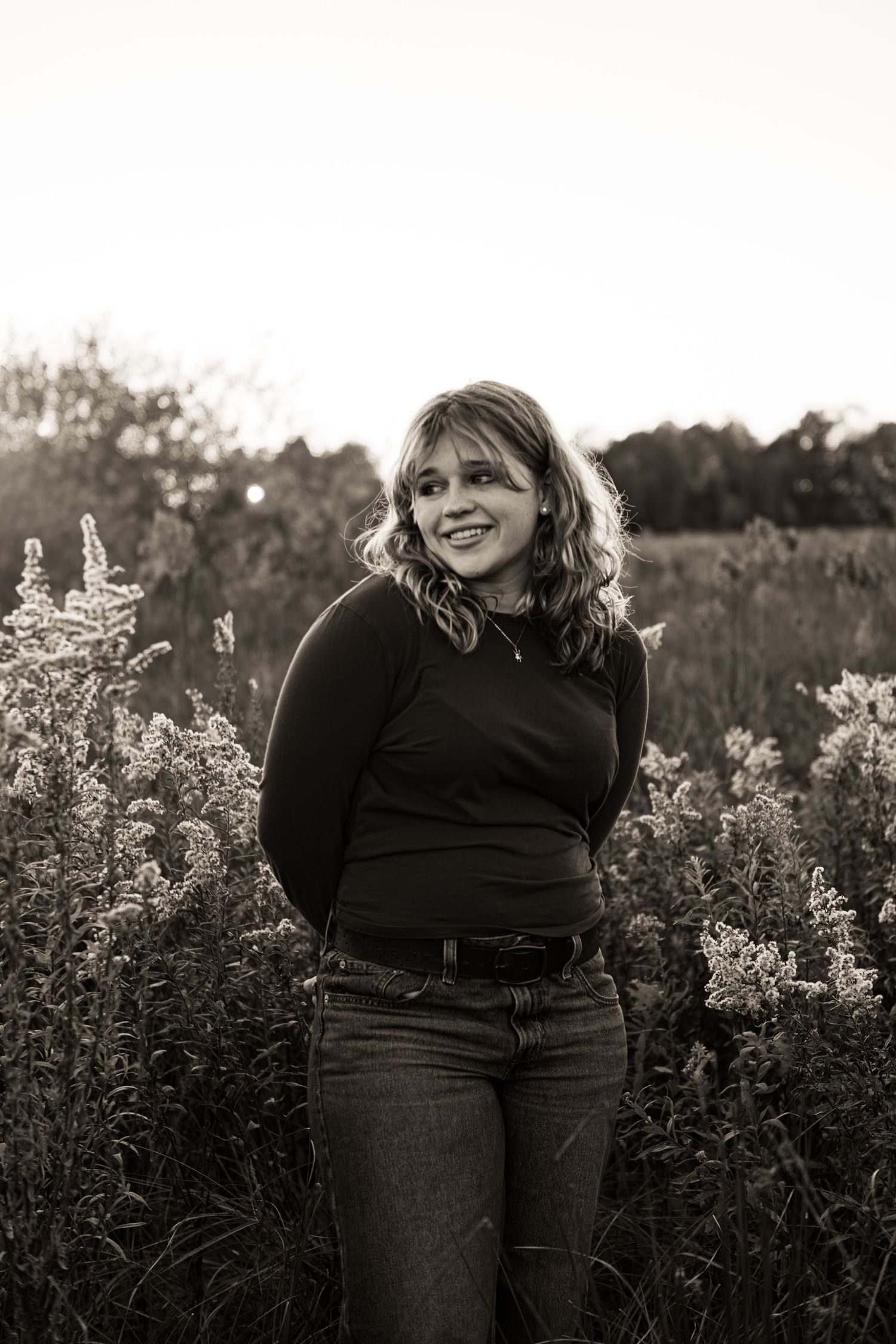 A person in black clothing stands in a field of wildflowers at sunset in a black and white photograph.
