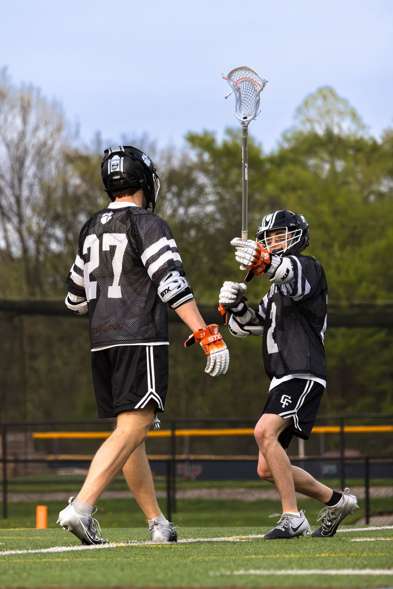 Two lacrosse players in black uniforms passing the ball during a match on a sunny day.