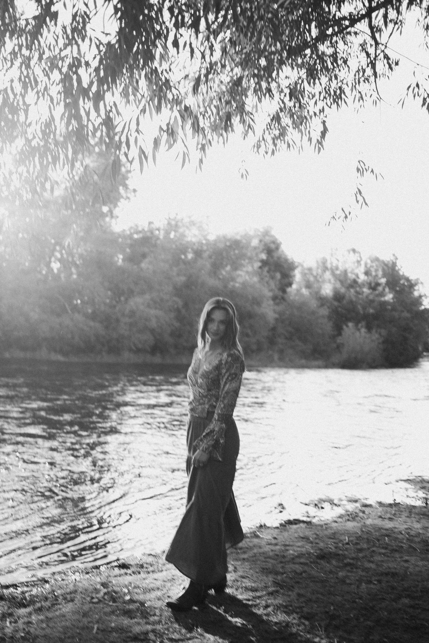 Black and white artistic portrait of a woman in a long skirt standing by a sunlit riverbank.