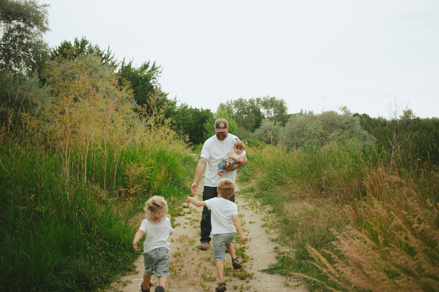 Father in white shirt walks sandy nature trail with two young blonde boys running ahead through green landscape.